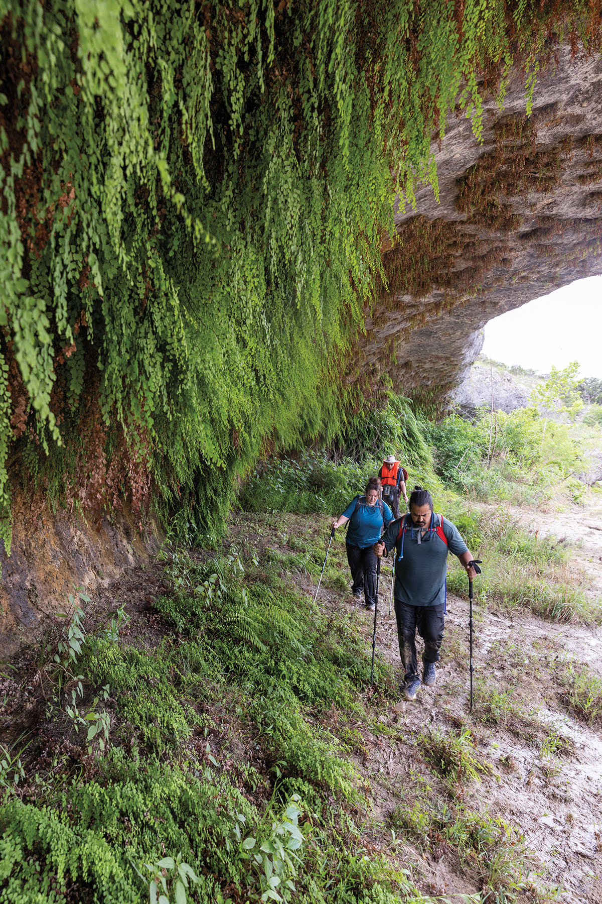 A group of people use hiking poles to traverse underneath a rock outcropping with moss