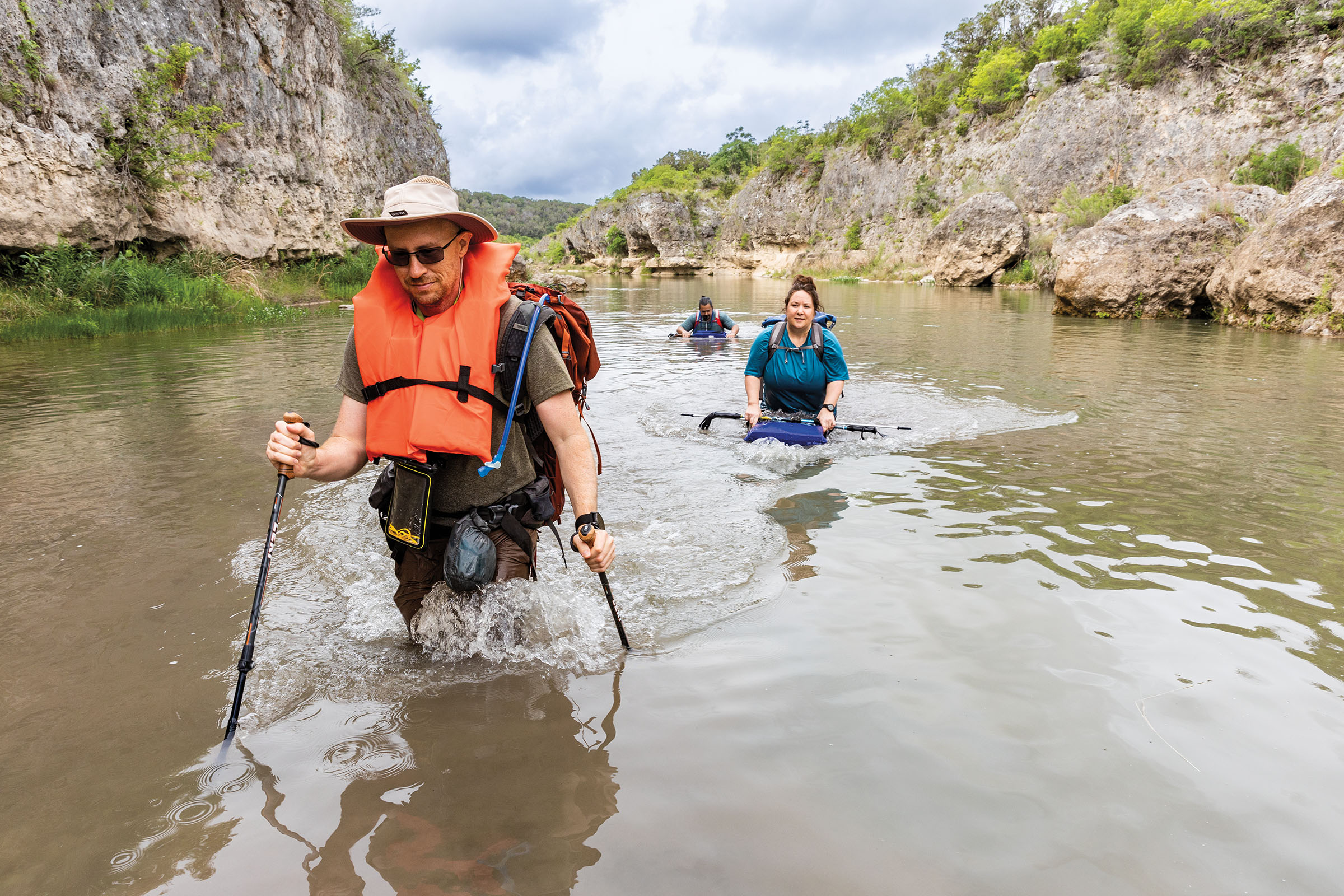 A group of hikers, one wearing an orange life vest, wade through murky water