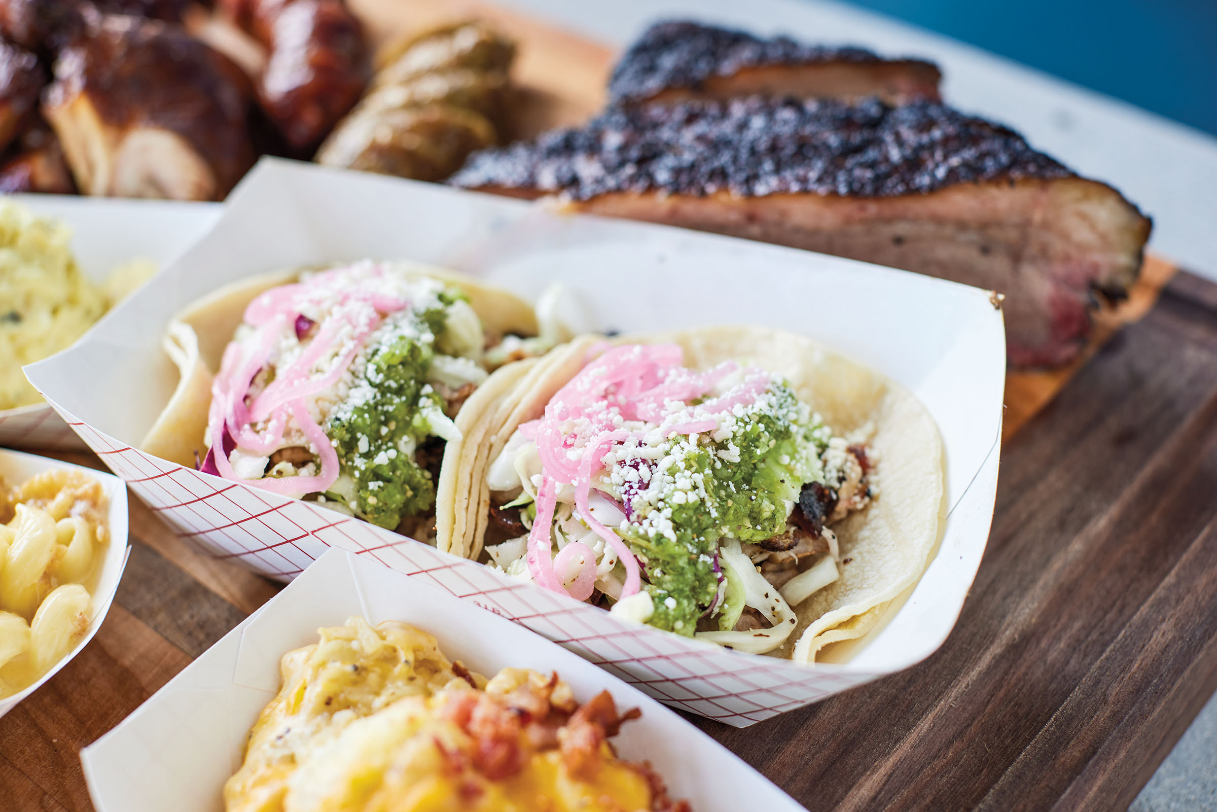 Two tacos with green salsa, pickled onions, cheese and meat served in a paper boat