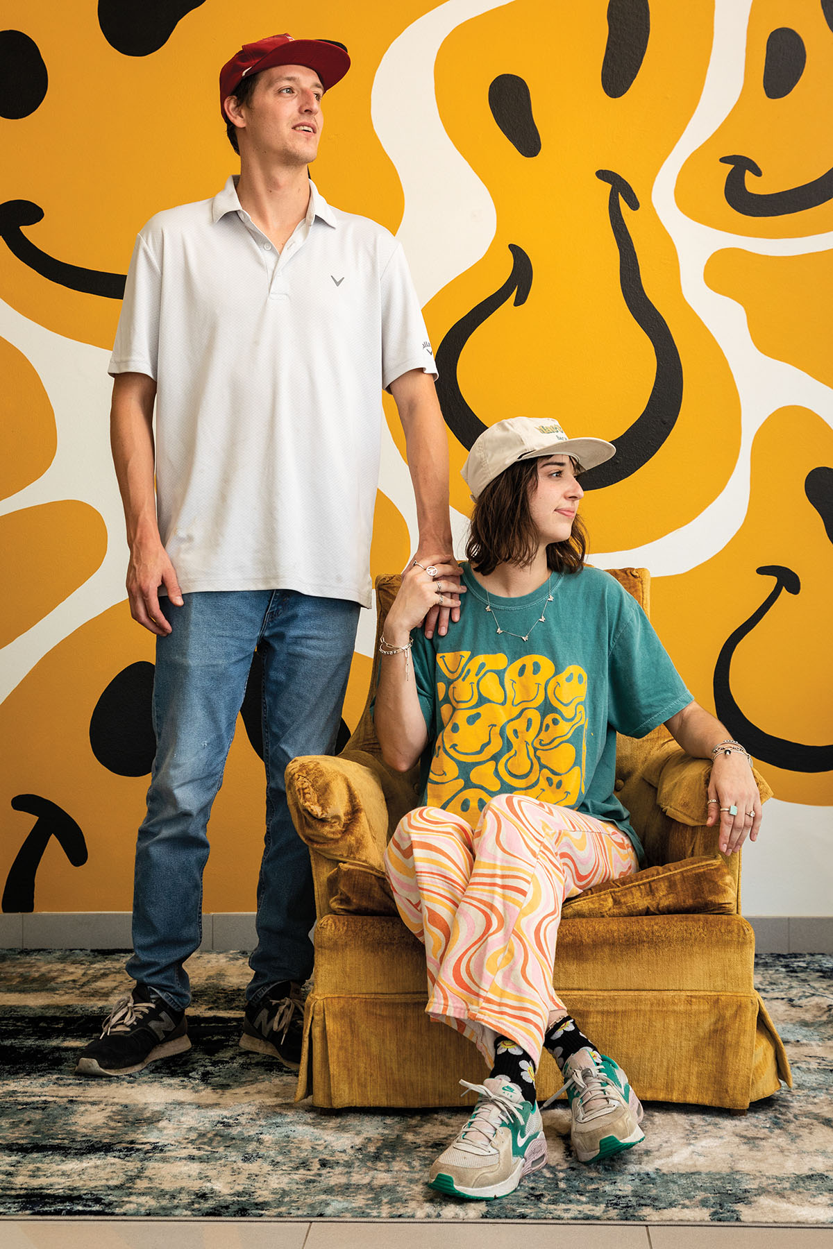 Two people wearing hats sit and stand in front of a wall painted with large yellow smiley faces