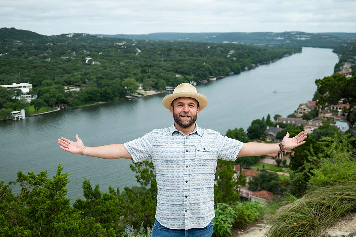 A man standing with his arms open wide with Lake Austin in the background.