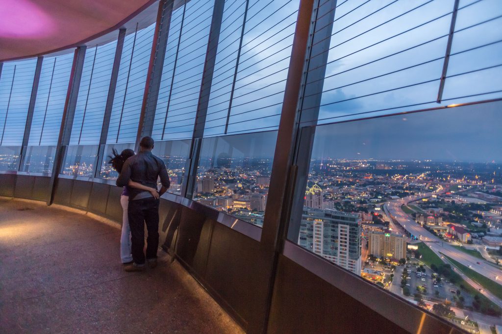 Two people stand next to large windows overlooking downtown San Antonio