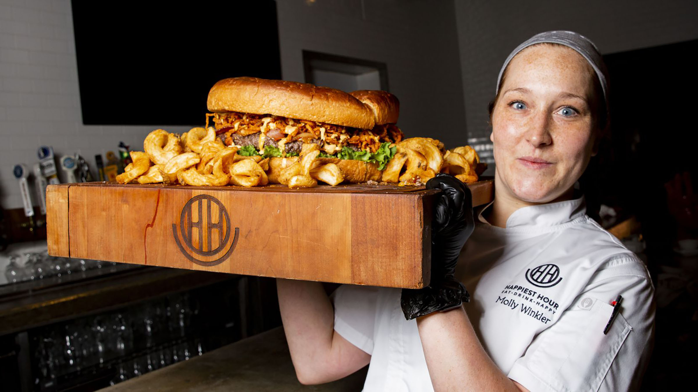 A person holds a large wooden cutting board with an oversize burger and large portion of fries