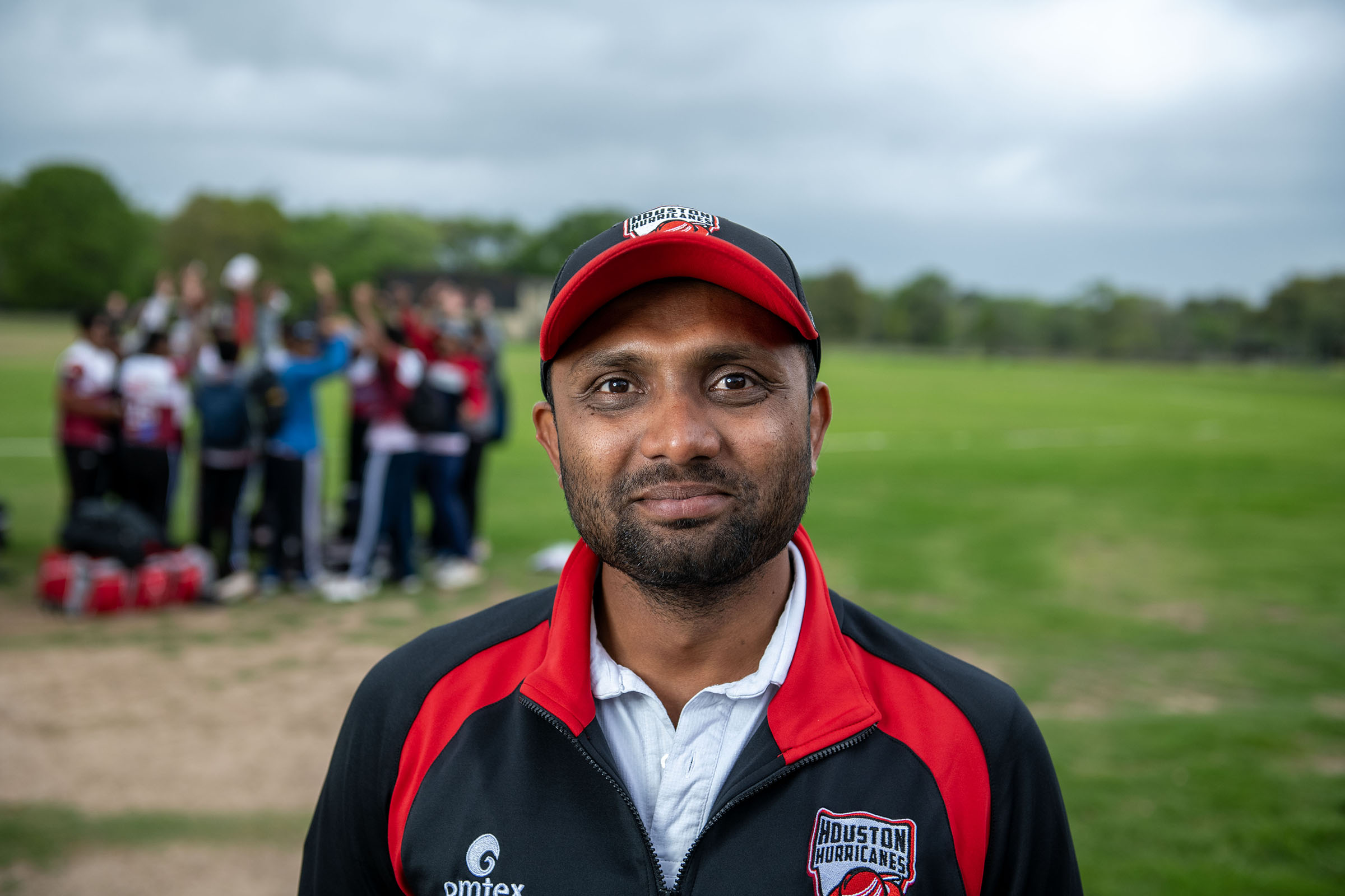 A man in a red and black cap and red and black athletic jacket stands on a cricket field 