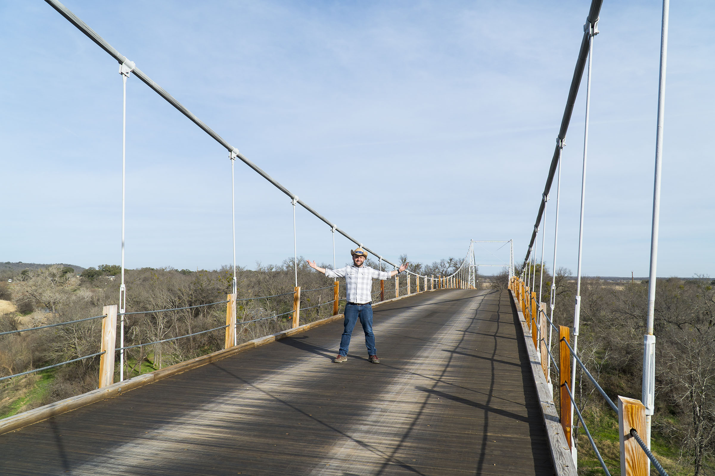 A man in a small cowboy hat stands on a swinging bridge raising his hands