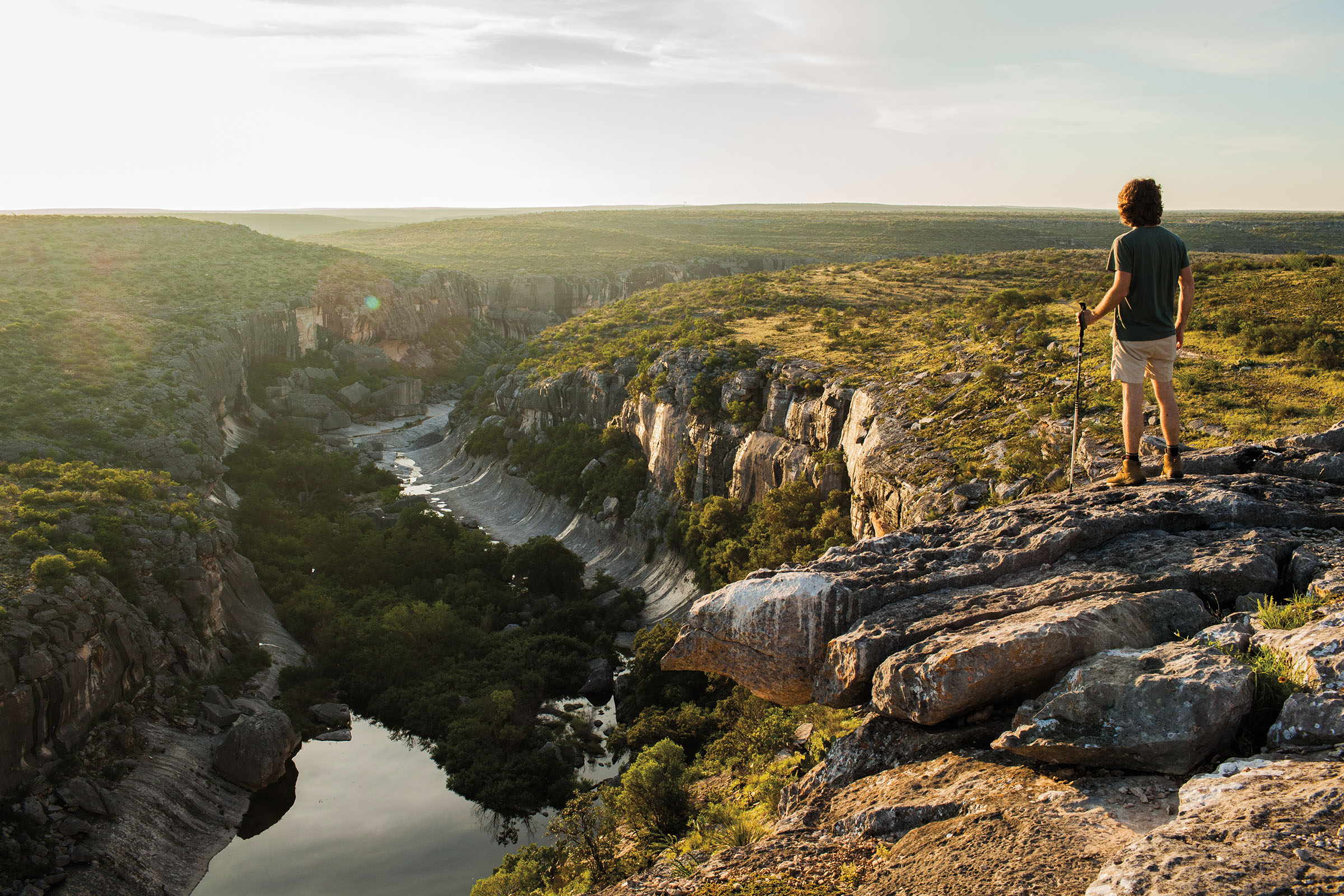 A person stands on a rock outcropping overlooking a placid river in a green scene