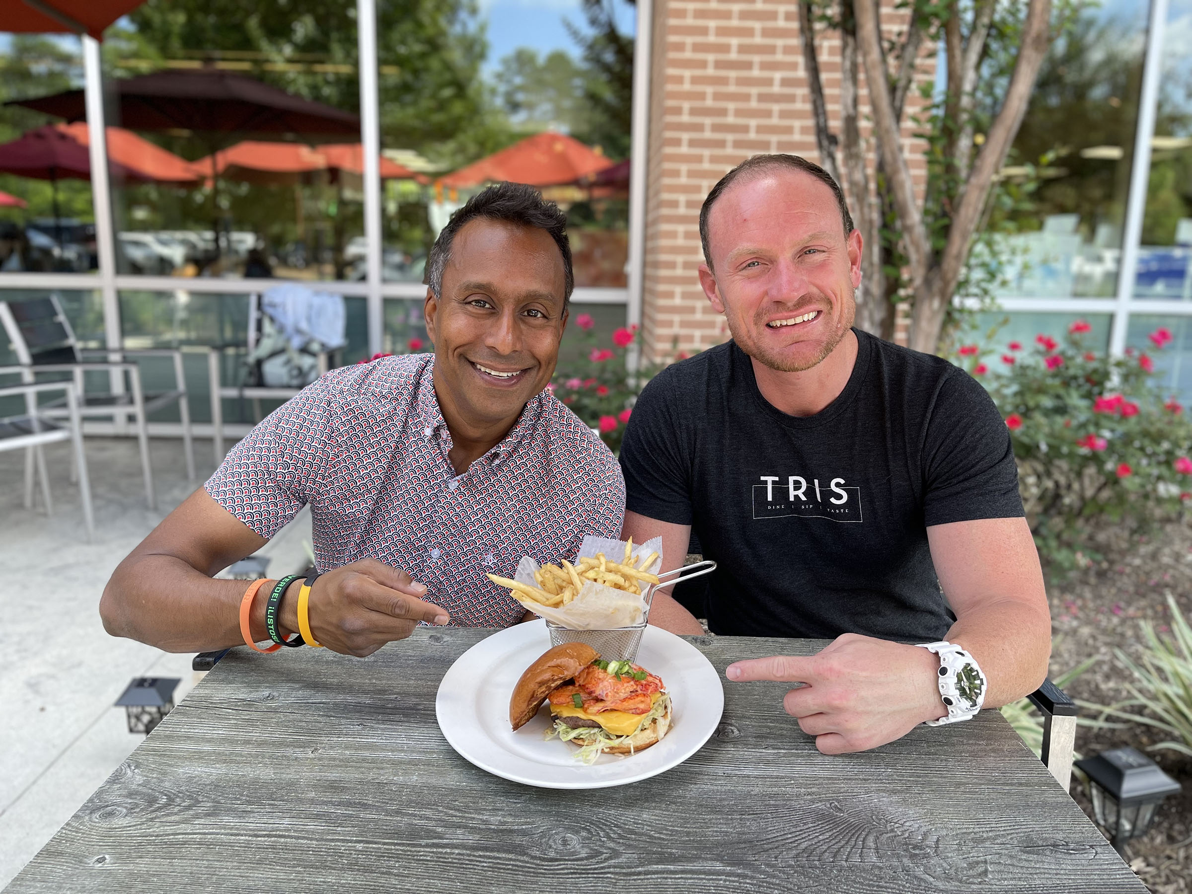 Two men sit at a table pointing at a burger topped with lobster and a side of fries