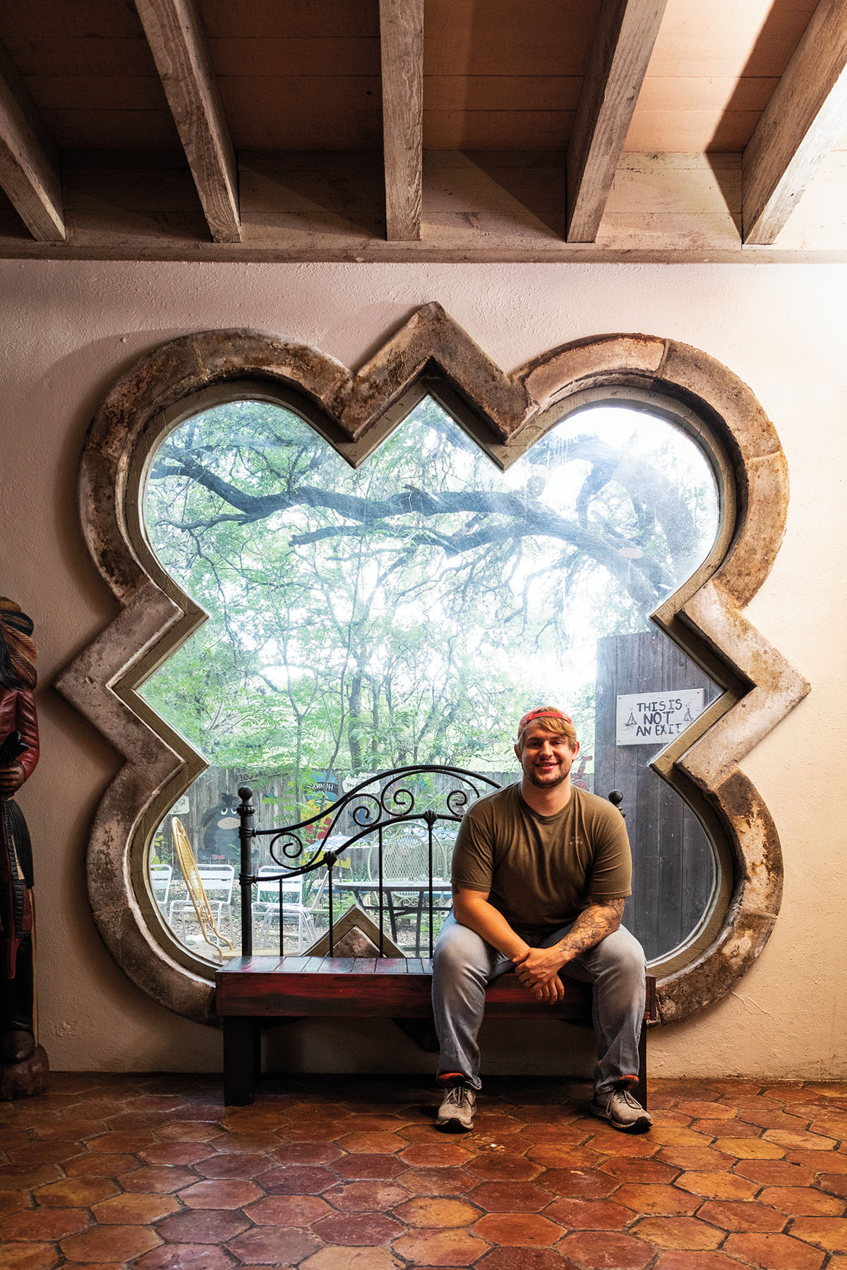 A man sits on a bench in front of a large window looking out toward live oaks