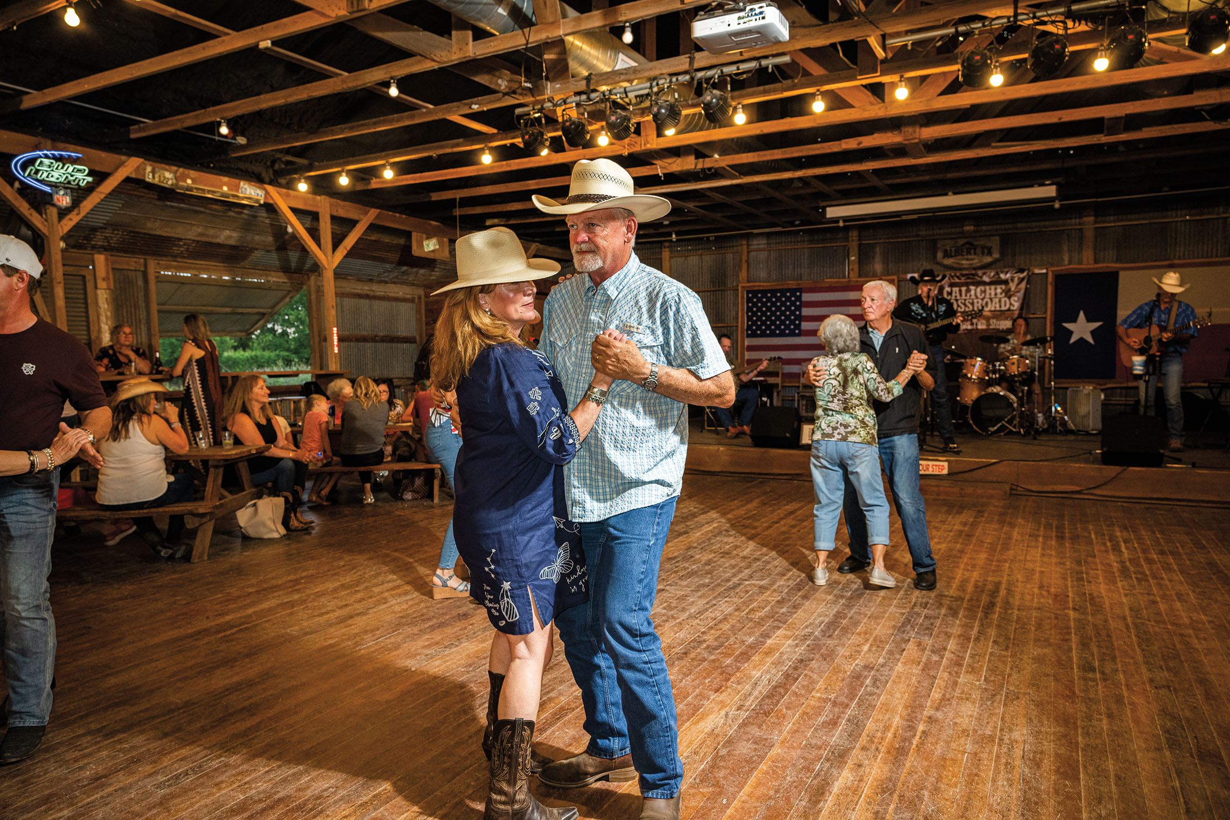 Two people wearing cowboy hats dance on a wooden floor inside a wooden dancehall