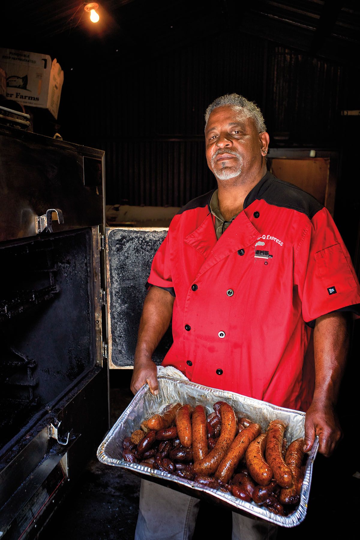 A man in a red shirt stands holding a large tray of sausage in front of a smoker