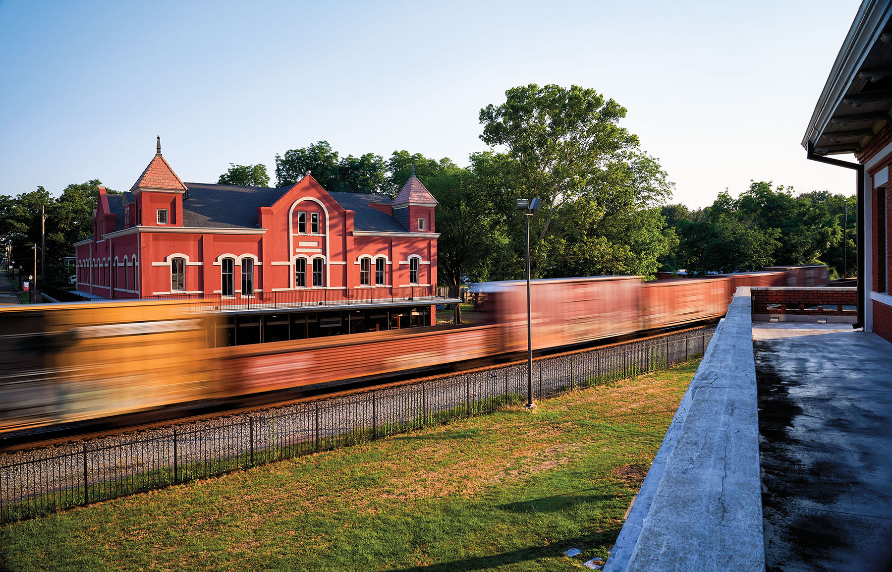 The blur of a freight train passes in front of a large red brick building with ornate rooftops