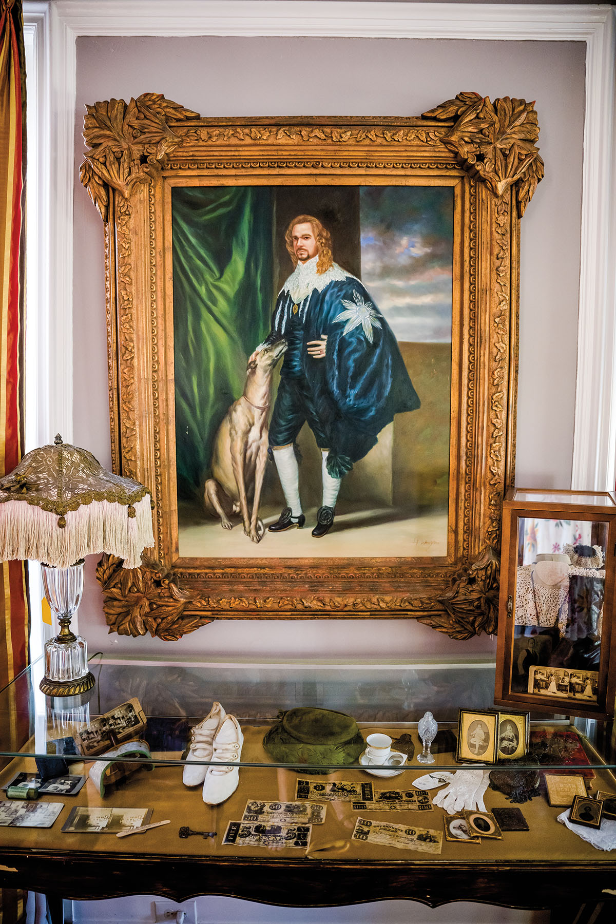 A framed portrait of a person wearing a navy cape petting a dog above a case of numerous antiques