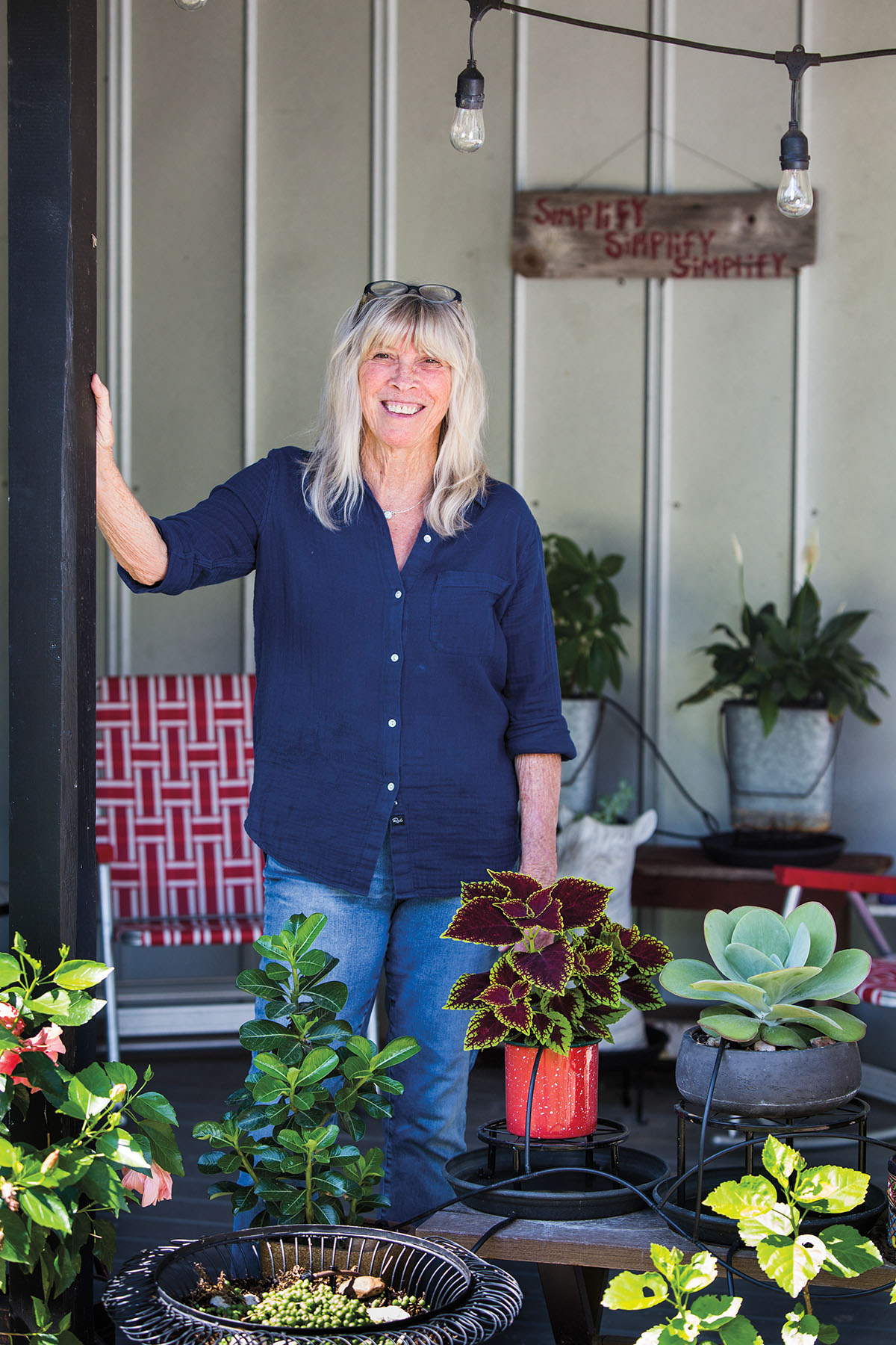 A woman in a blue shirt stands among several plants outside of a house