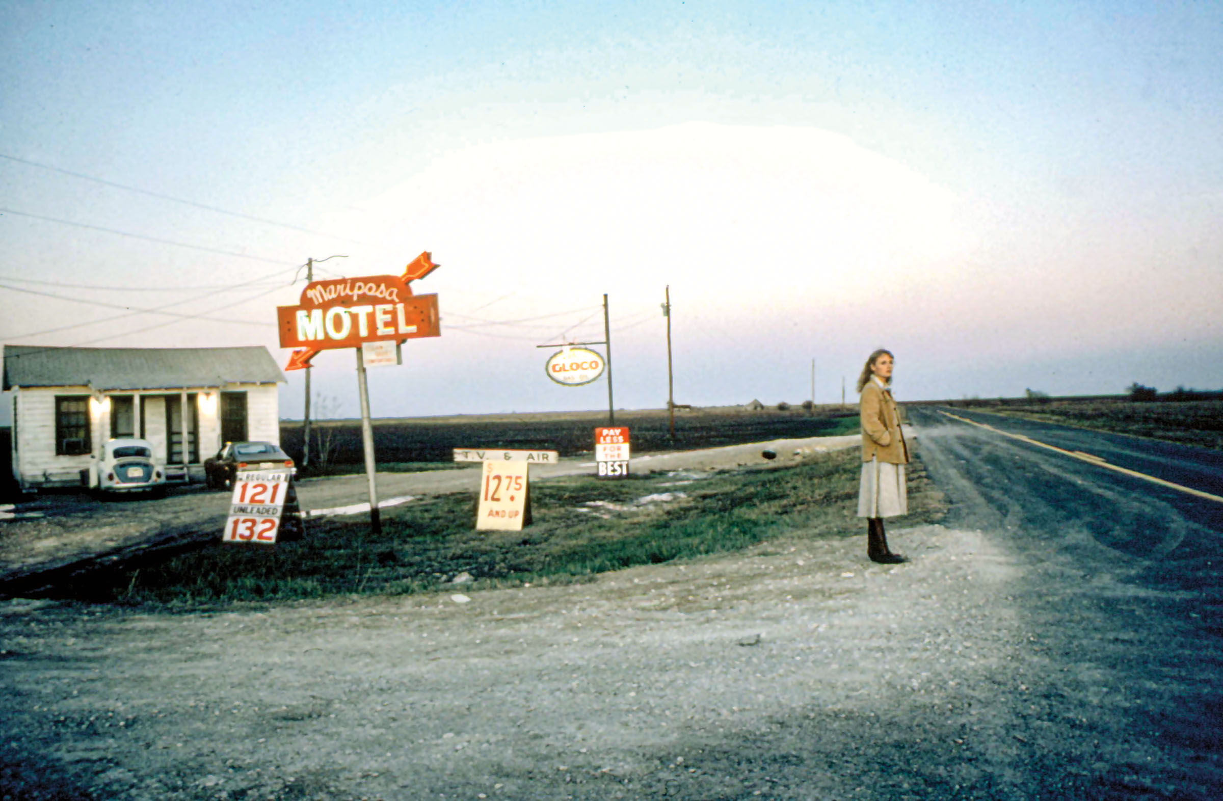 An image of a woman standing along a dirt road next to a sign reading 'Mariposa Motel'