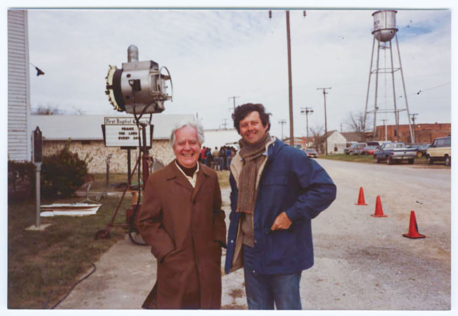 Two men in jackets stand smiling on a dirt road in a town