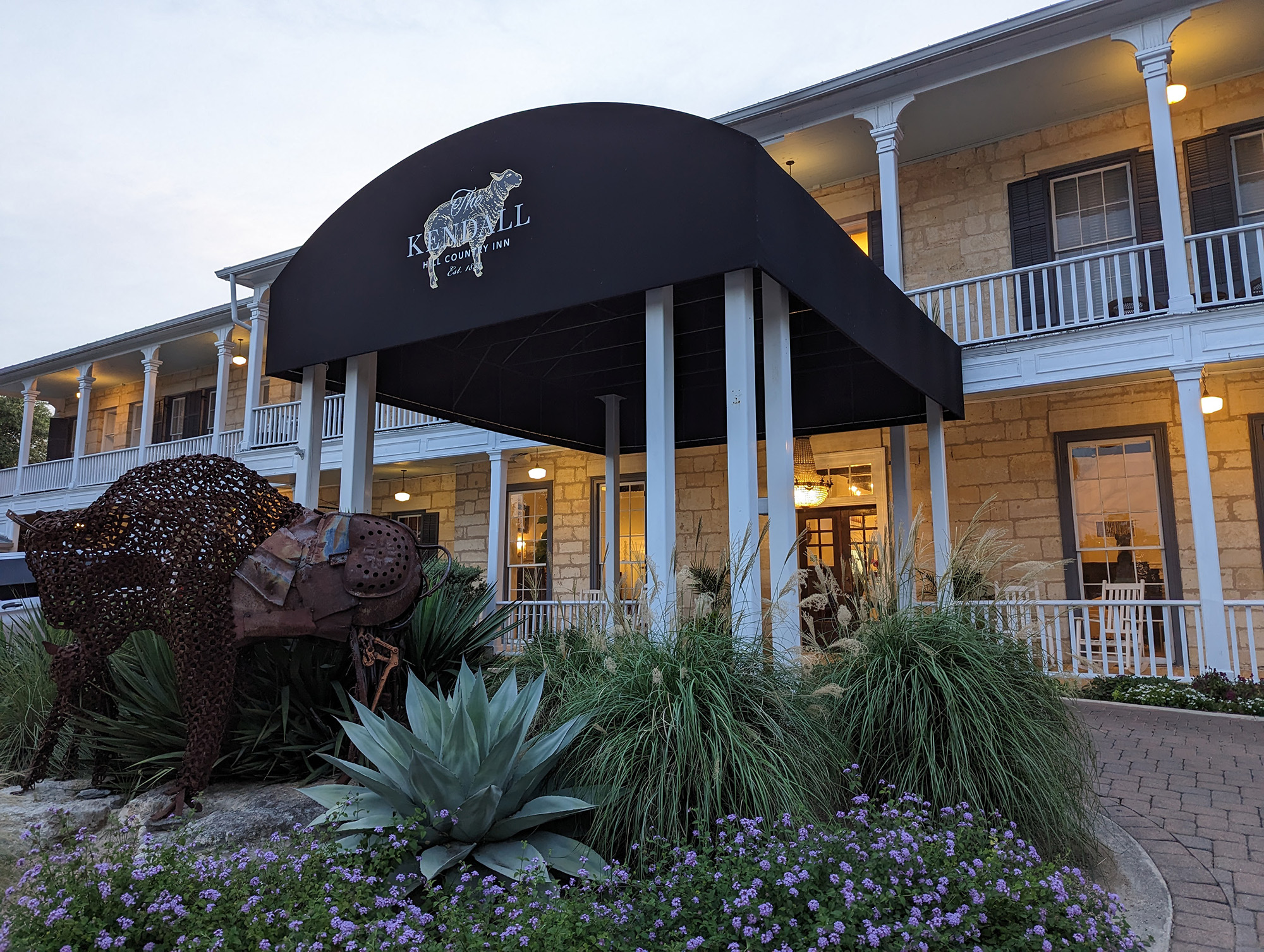 The front entrance to The Kendall, a hotel painted white with a black-and-white portico and a logo featuring a sheep with the words "The Kendall Hill Country Inn Est. 1859." A rusted metal statue of a buffalo is in front of the entrance.