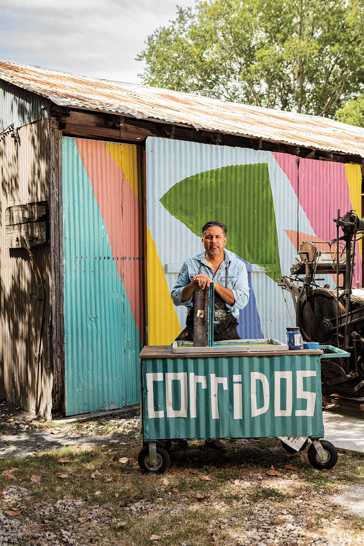 A man stands behind a teal metal sign reading "Corridos"
