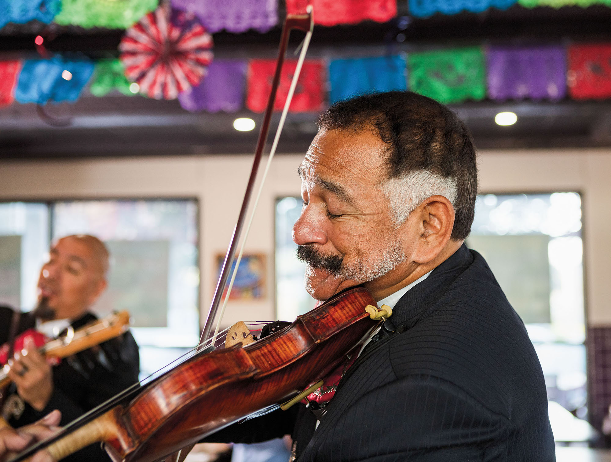 A man plays a rich wooden guitar in front of other performers and brightly-colored papel pacado flags