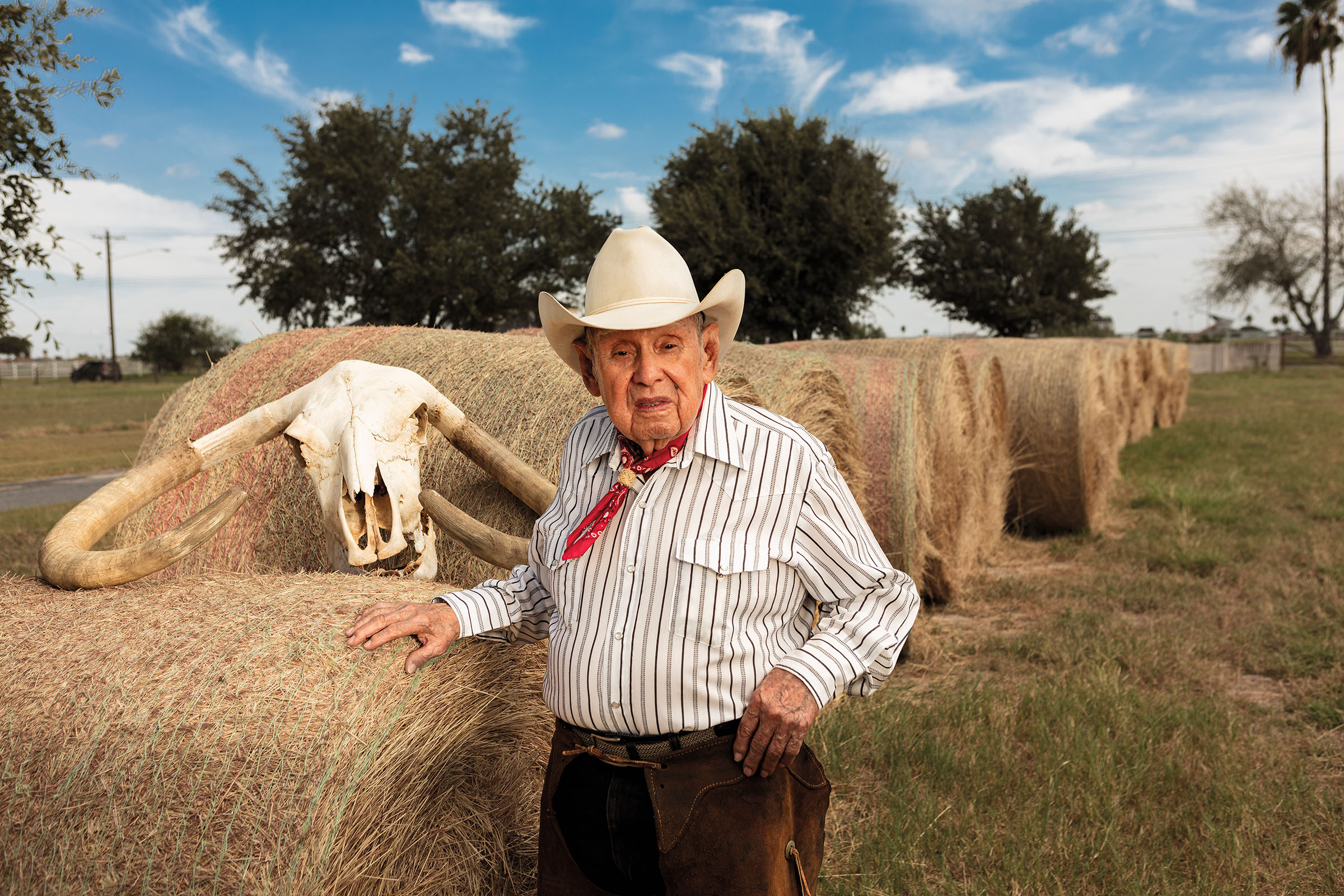 A sharply-dressed man in a white cowboy hat stands in front of large bales of hay and a cow's skull