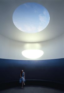 A woman looks up at a skylight to blue sky with the reflection of sunlight on the wall behind