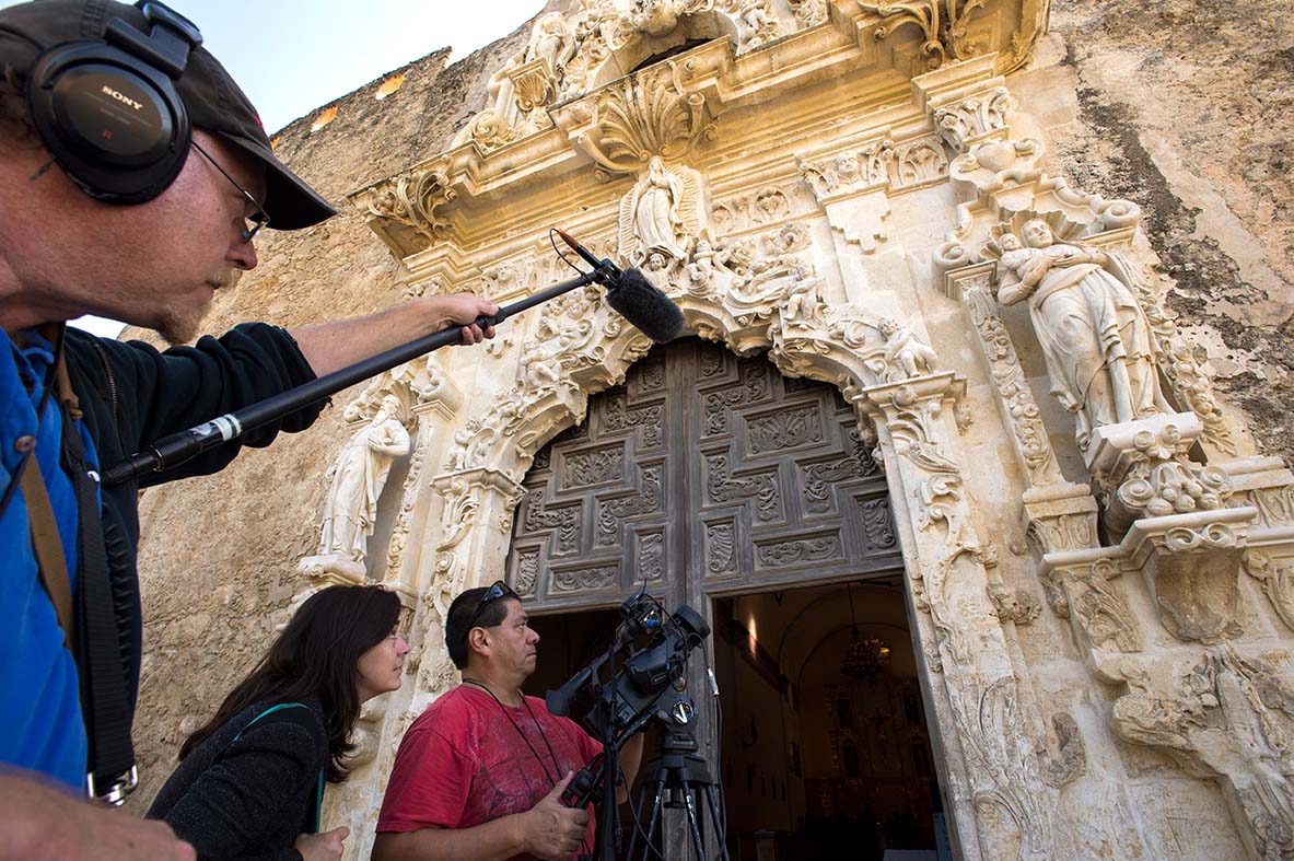 A man holds a boom mic while another films at a historic structure.