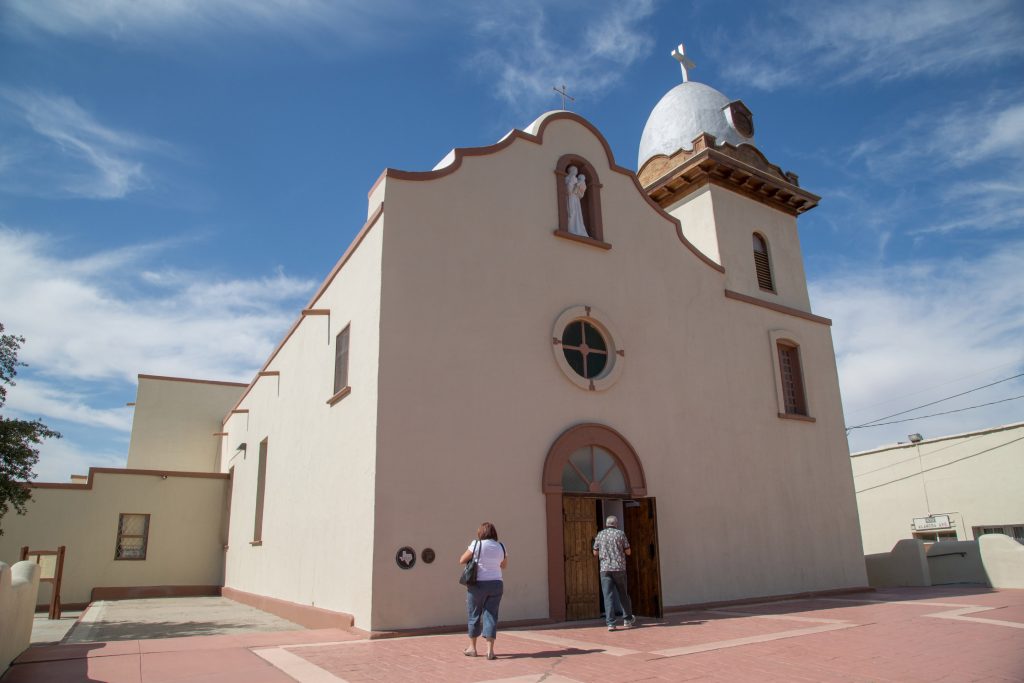 Two individuals stand outside of a whitewashed adobe building with a steeple.
