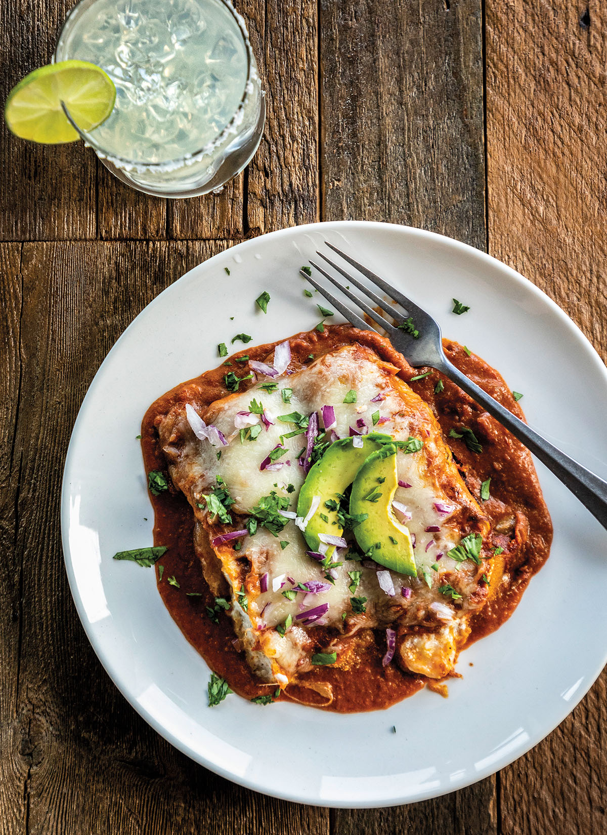An overhead view of bright red enchiladas topped with red onion and avocado, paired with a margarita