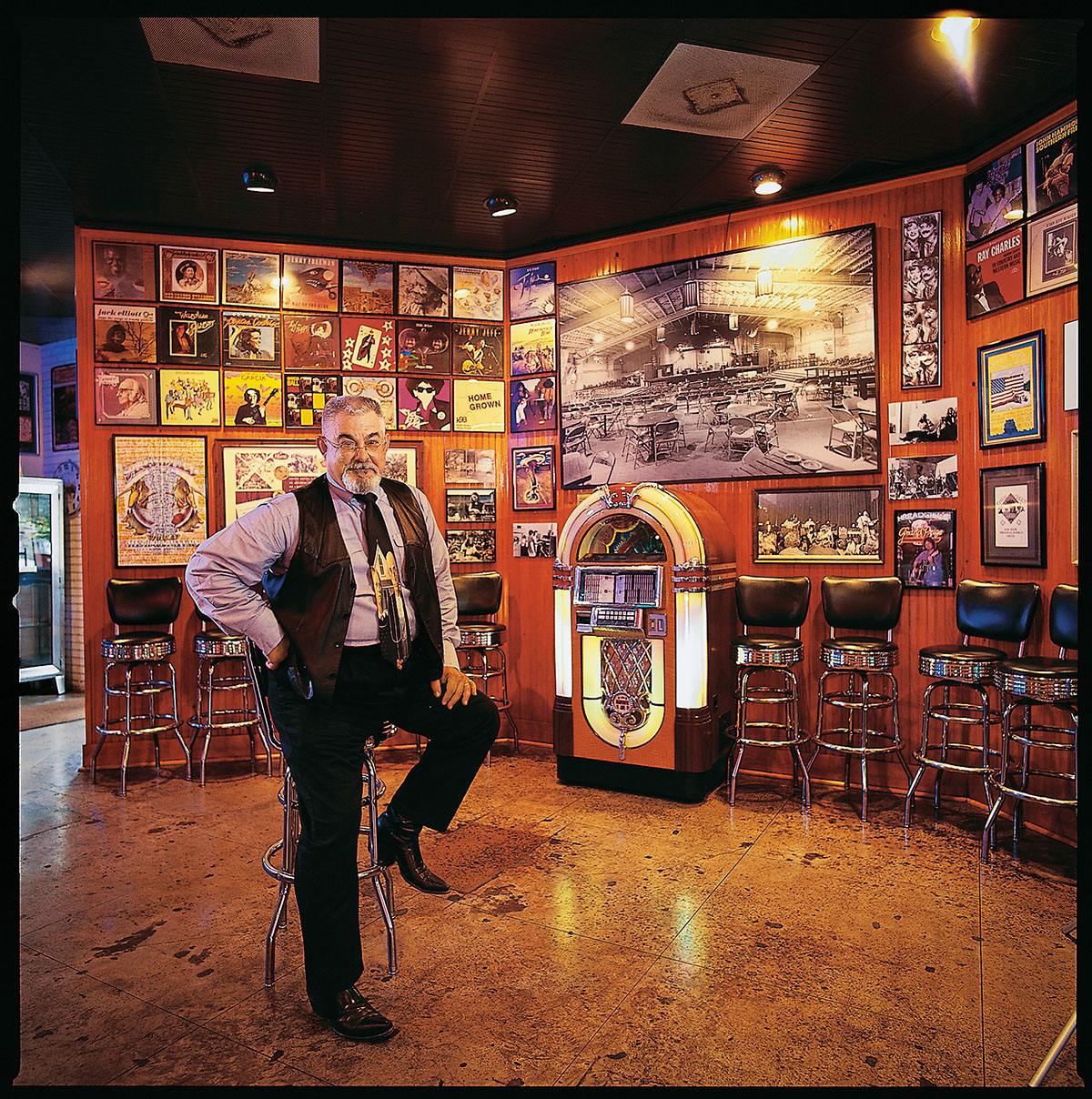 A man in old Western wear stands with his leg propped on a stool inside of a richly painted room decorated with music memorabilia