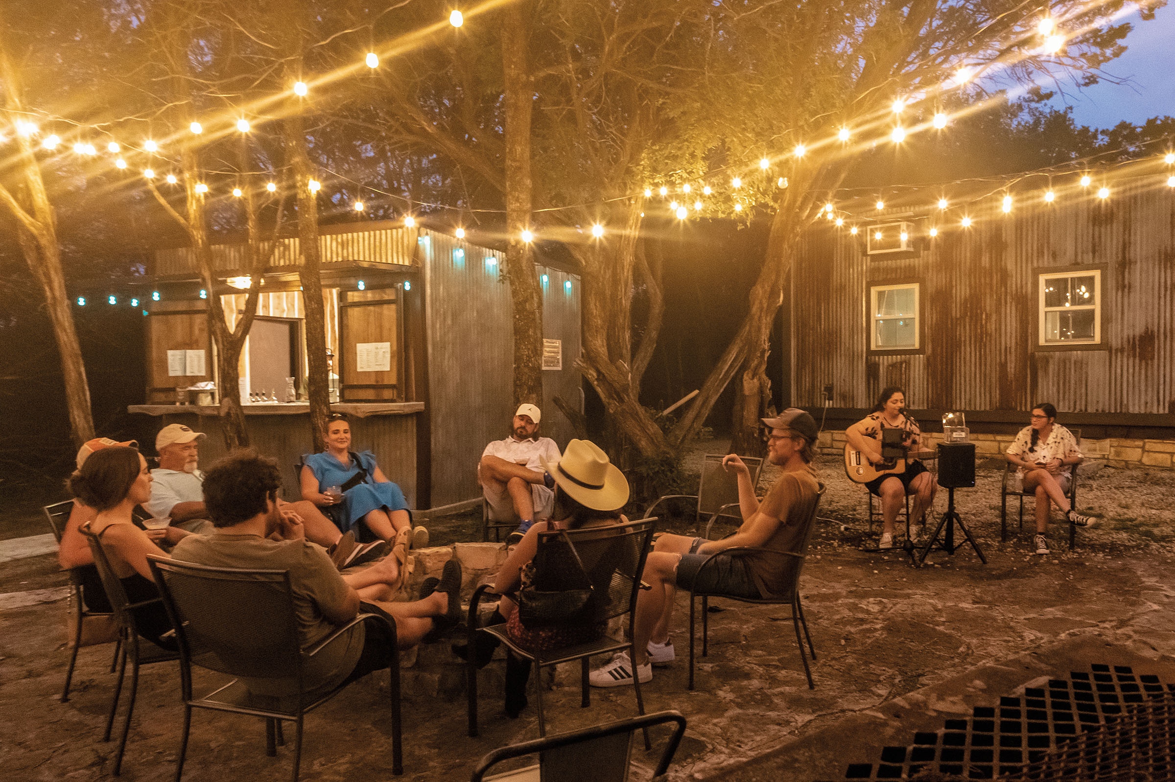 String lights hanging with people sitting in chairs outside.