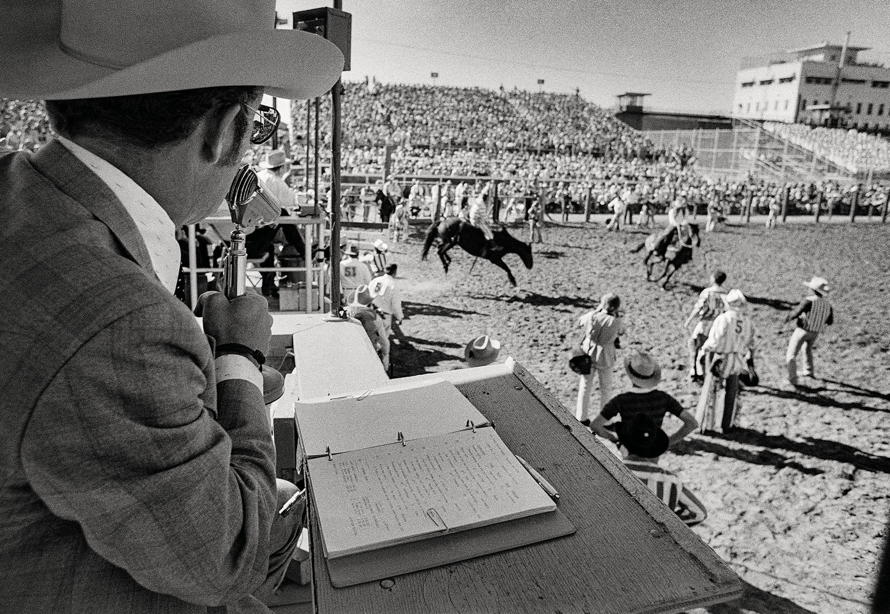 A man wearing a cowboy hat looks down over a dirt field of cowboys and wranglers participating in a rodeo