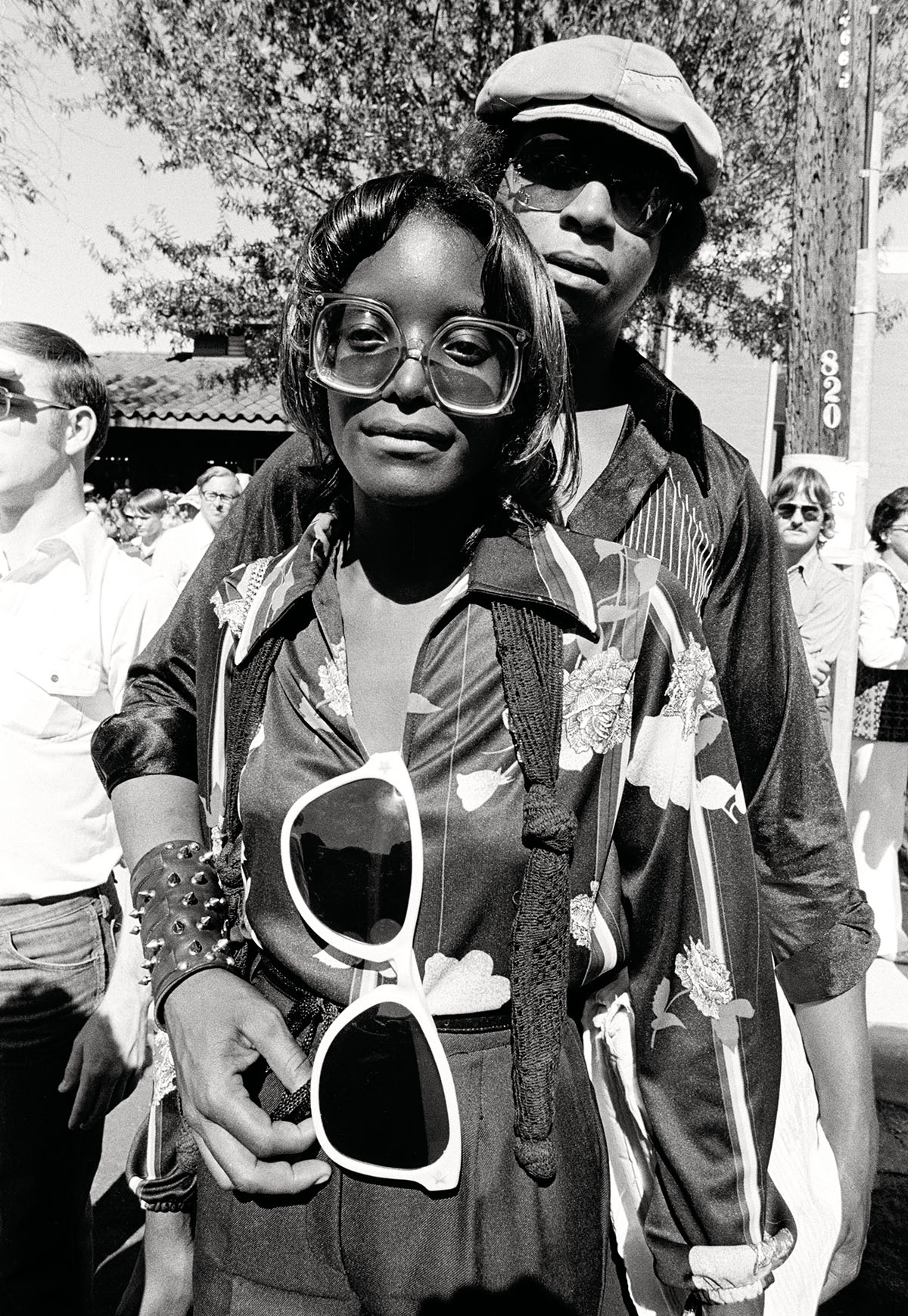 Two people dressed in typical 70s attire stand posting for the camera. The front woman wears large sunglasses around her neck