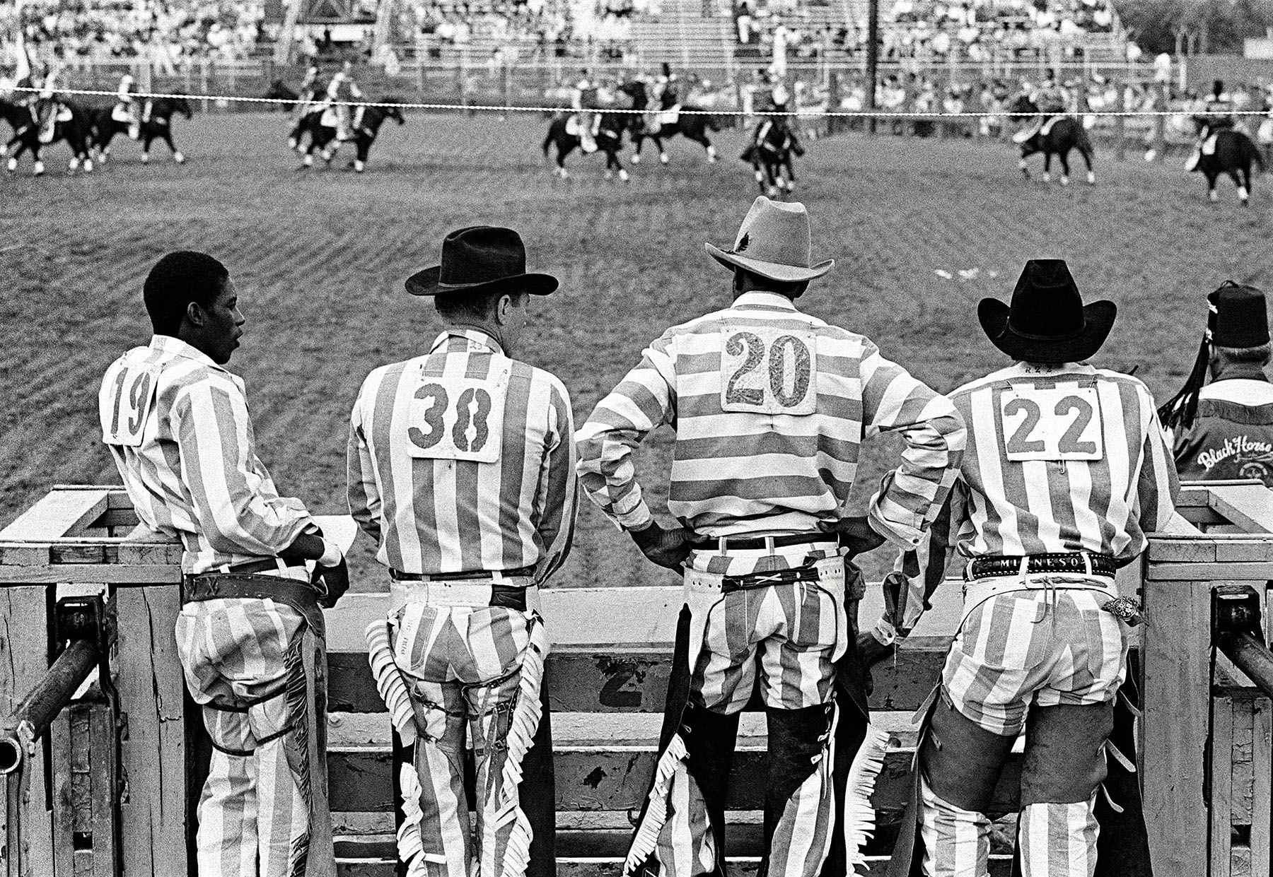 A group of people in striped jumpsuits and cowboy hats stand along a fence