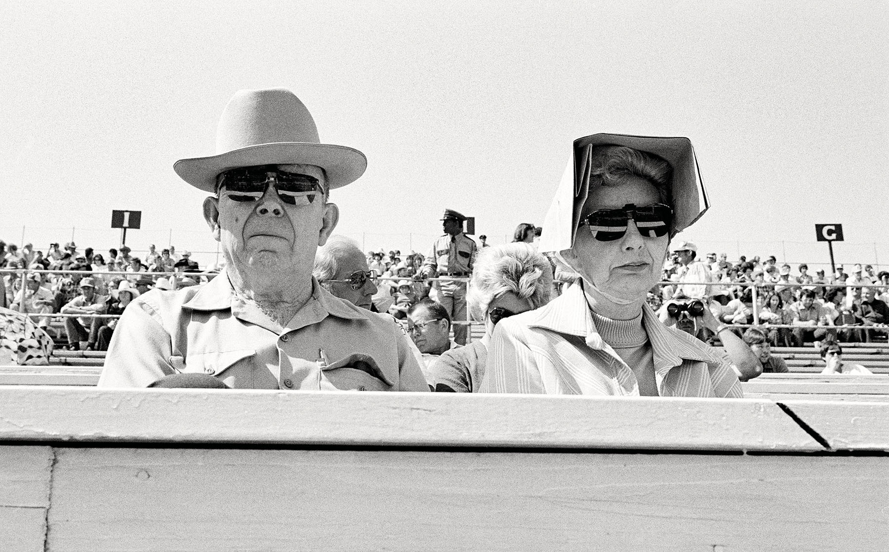 Two older people sit wearing sunglasses in the stands of a rodeo among other people