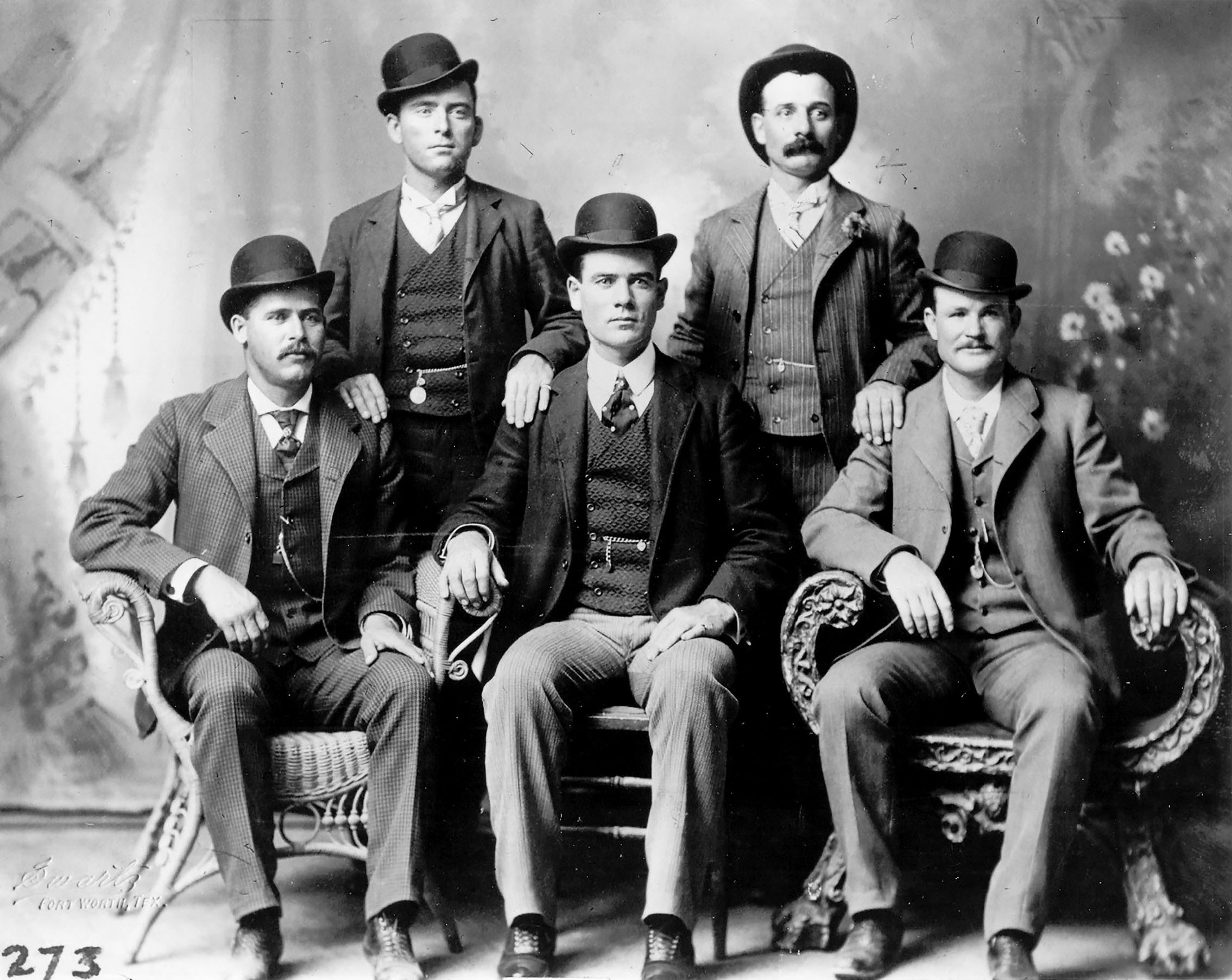 A black and white photograph of a group of men sitting on a bench wearing suits and hats