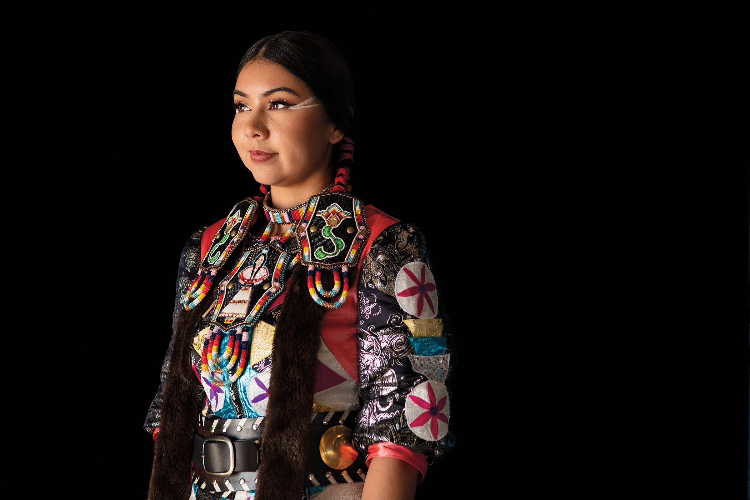 A woman dressed in brightly-colored clothes and colorful beaded headwear poses on a black background