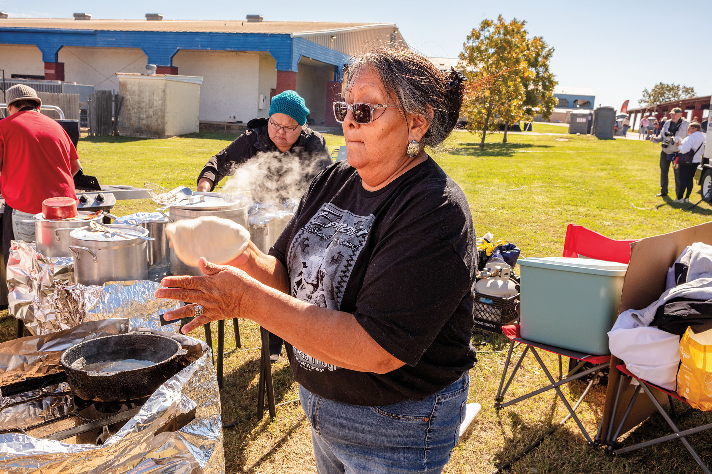 A woman in a black t-shirt holds a small piece of tan fry bread while others cook behind and around her in an outdoor setting