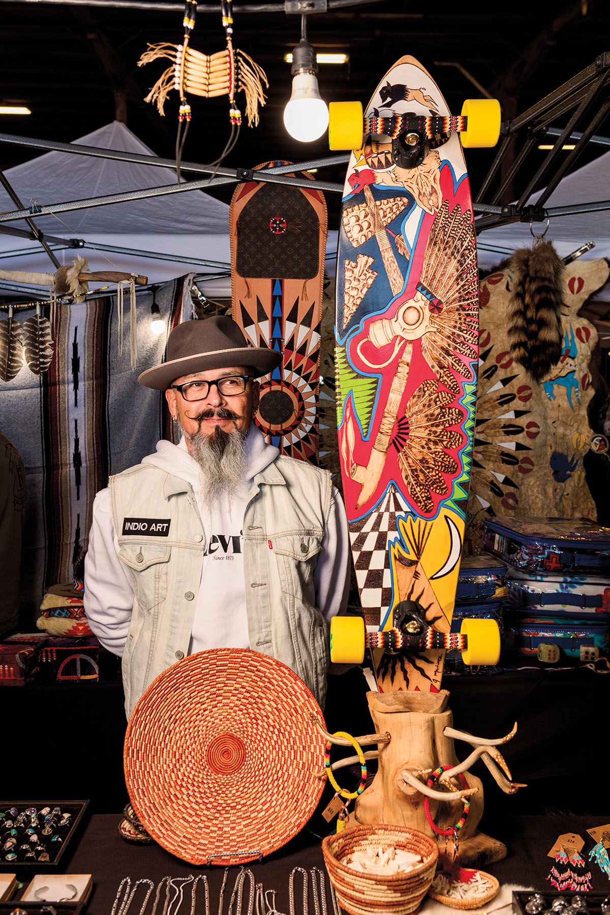 A man with a hat and long beard stands next to a brightly-painted skateboard