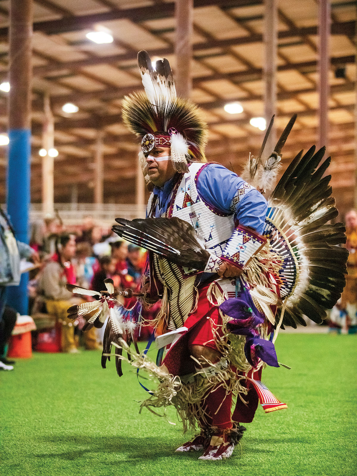 A man in ornate dress with numerous beads and feathers dances on green grass inside of a wooden building