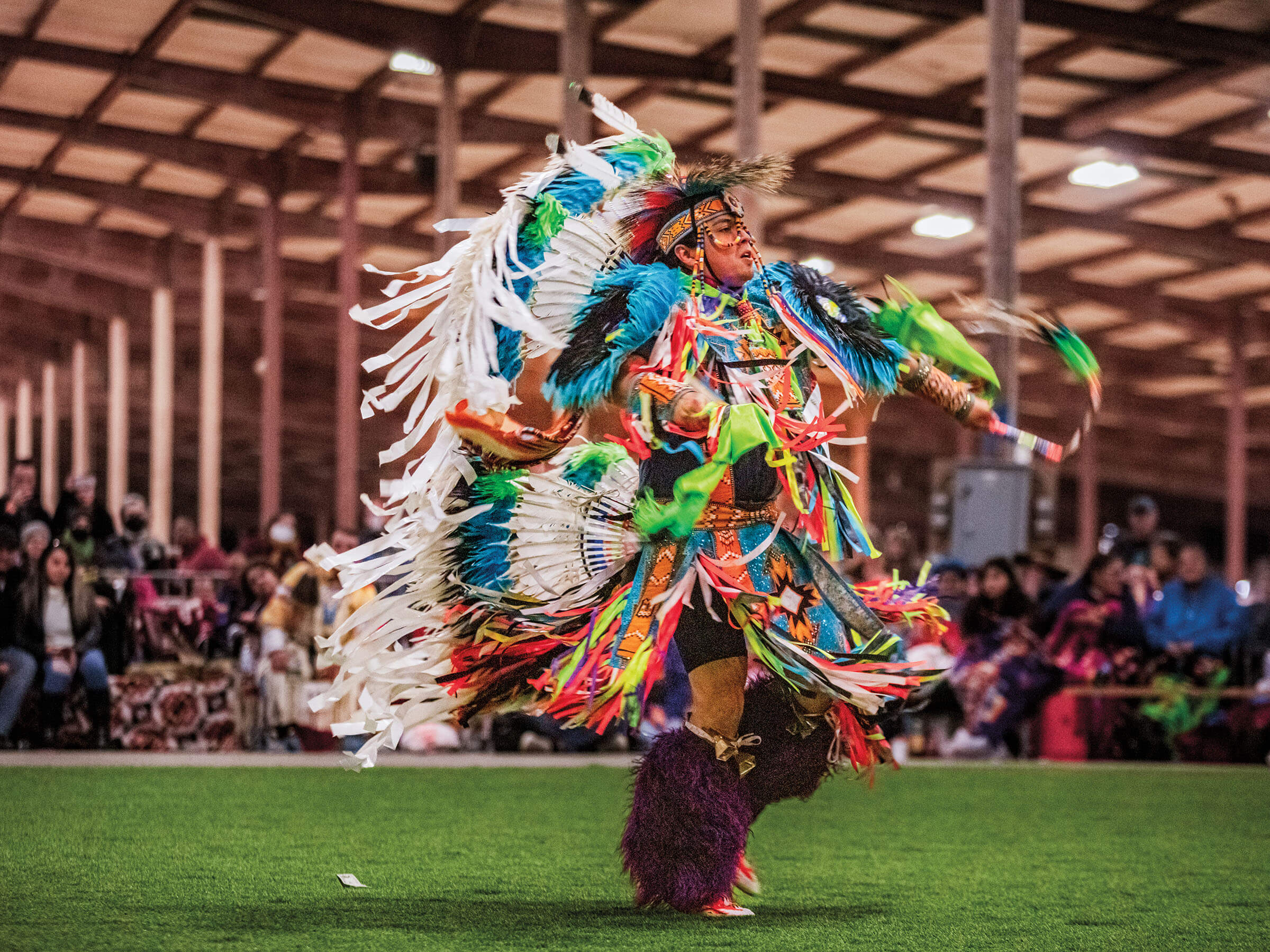 A person in brightly-colored clothing with numerous ribbons and feathers dances on a green grass area