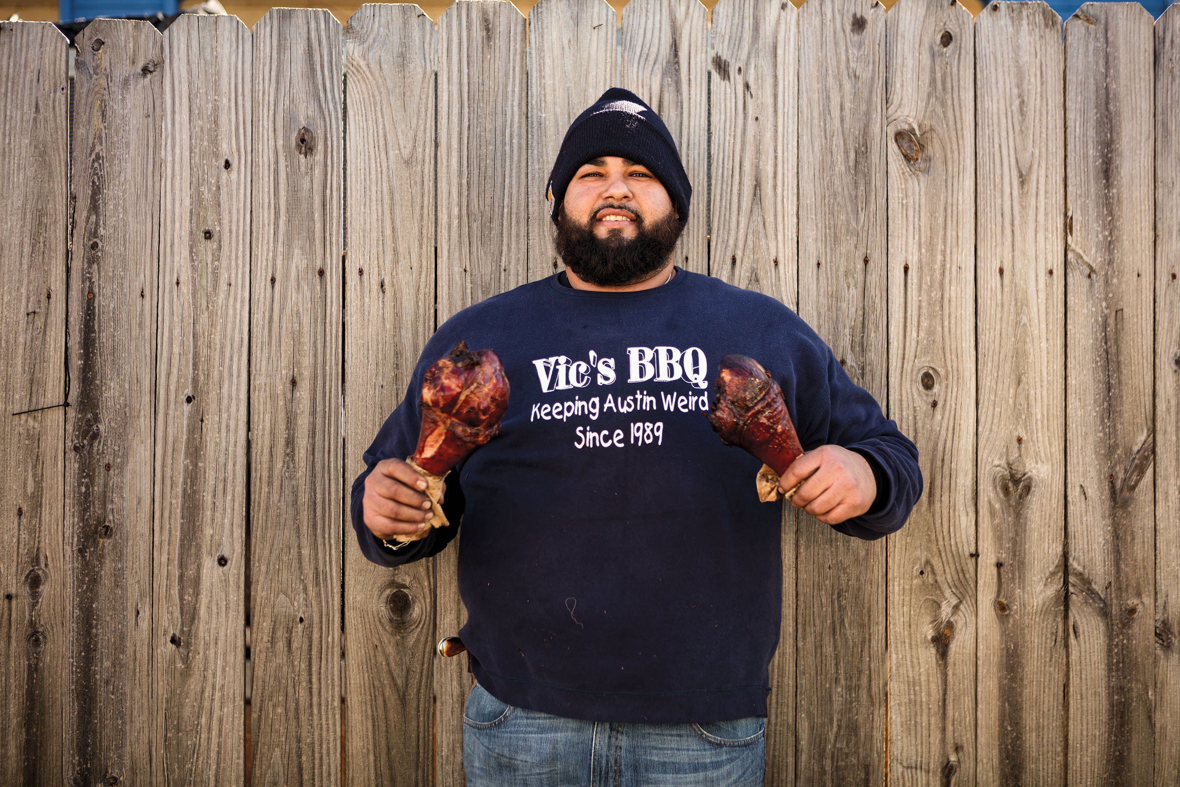 A man wearing a blue sweatshirt reading "Vic's BBQ" poses with two large turkey legs, one in each hand