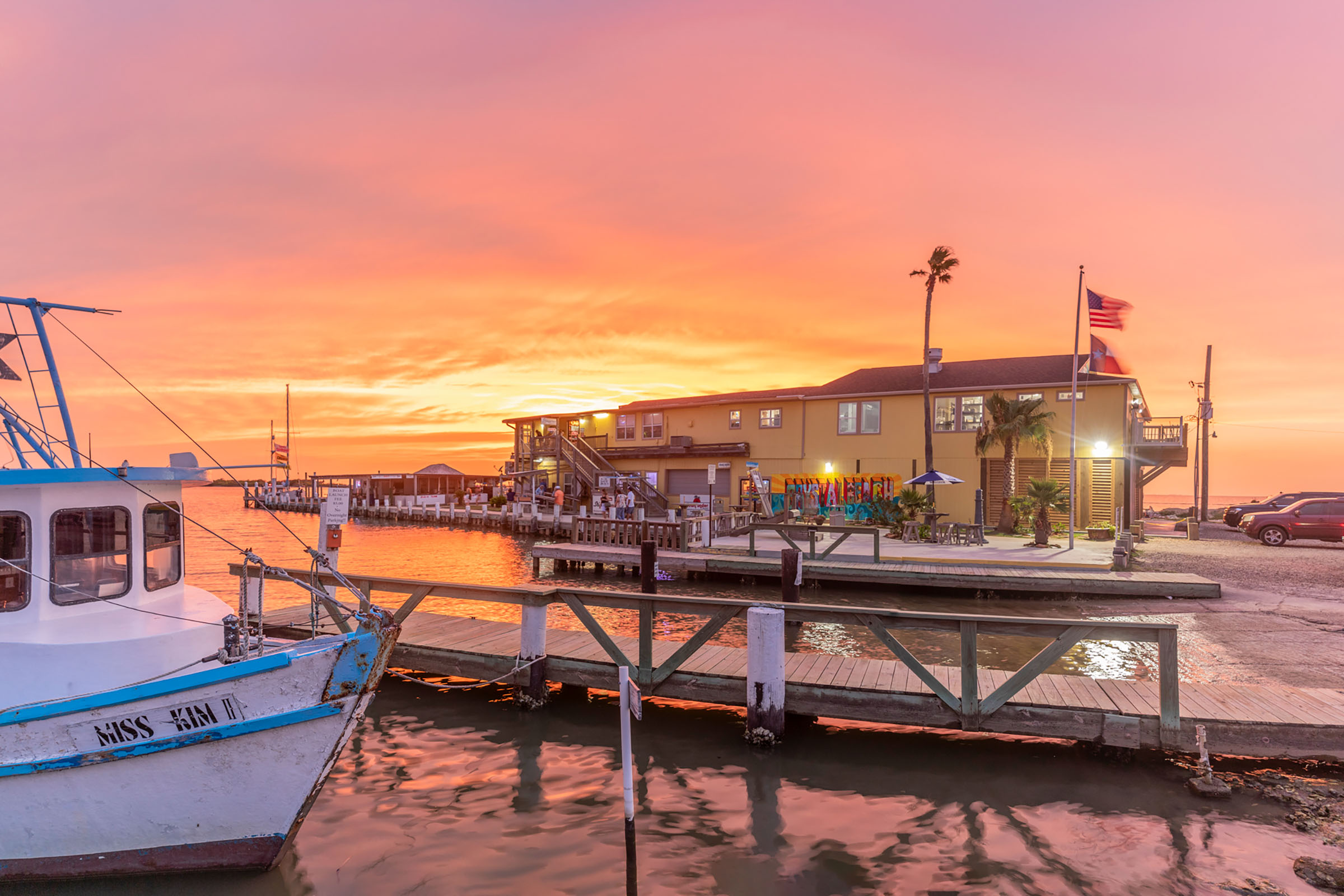 A wide shot of a building and boat on the water with a sunset in the background