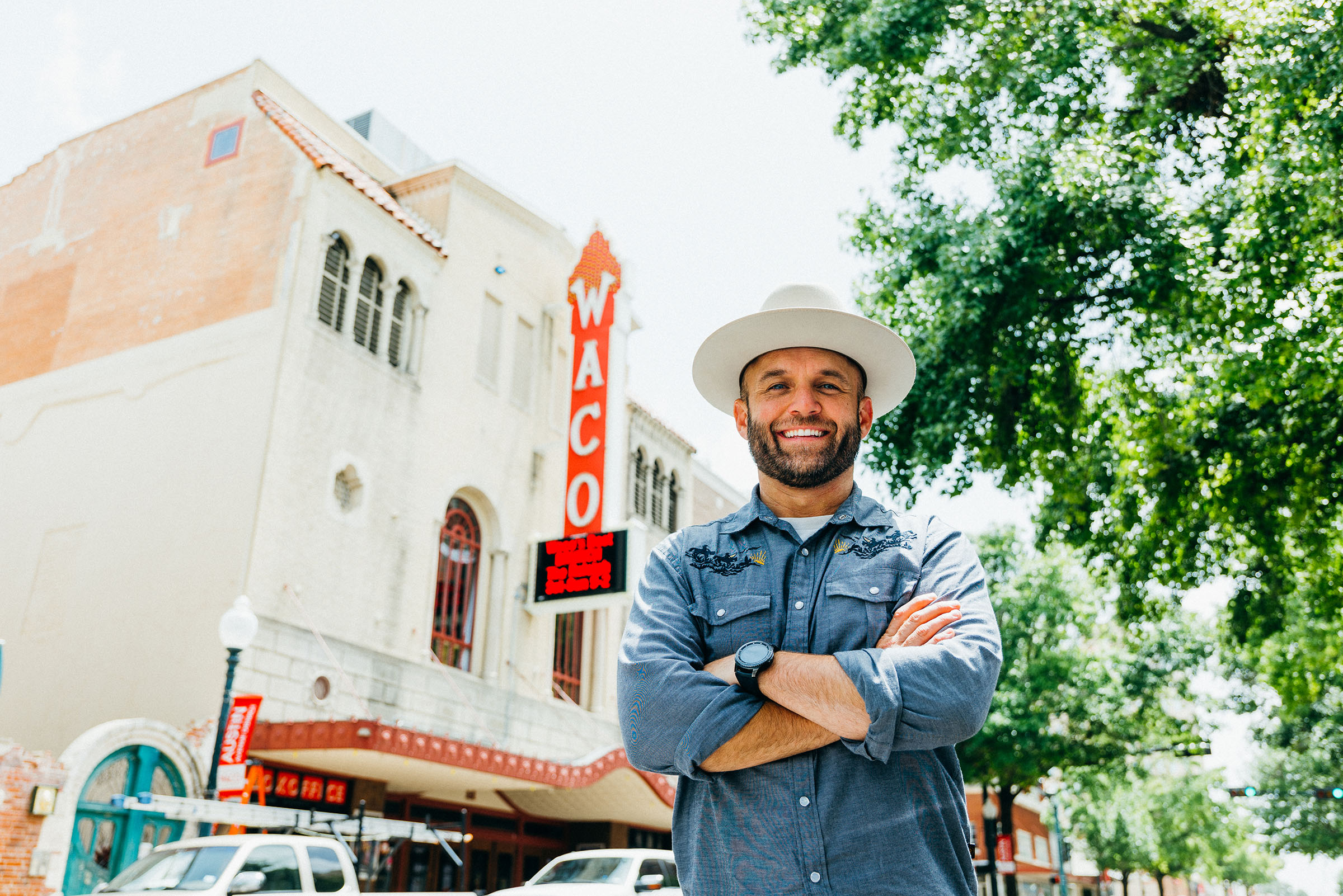 A man in a blue button-down shirt and small cowboy hat stands with his arms crossed in front of a theater with a red "Waco" sign on the front