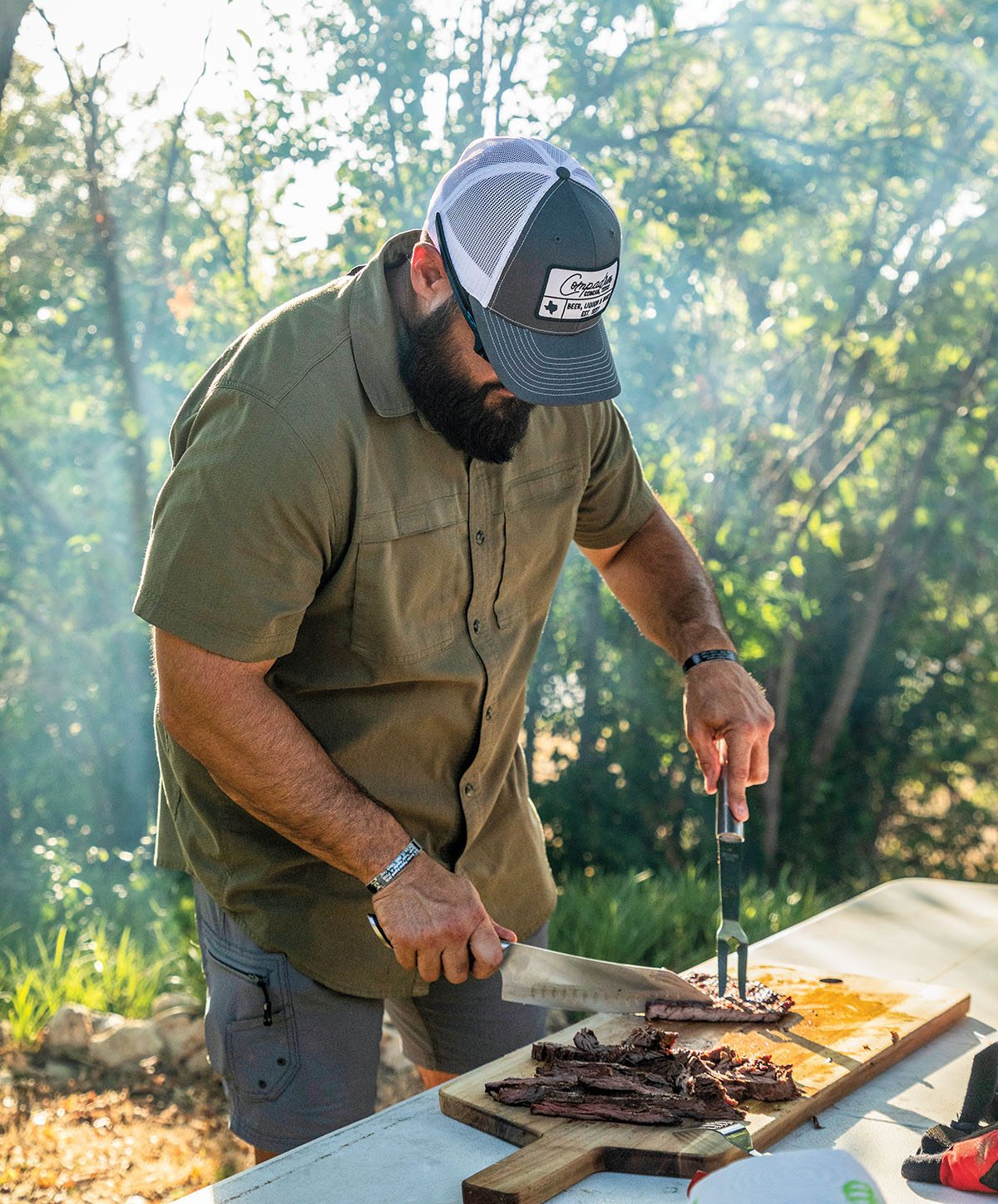 A man in a ball cap and green shirt cuts meat on a platter in an outdoor setting