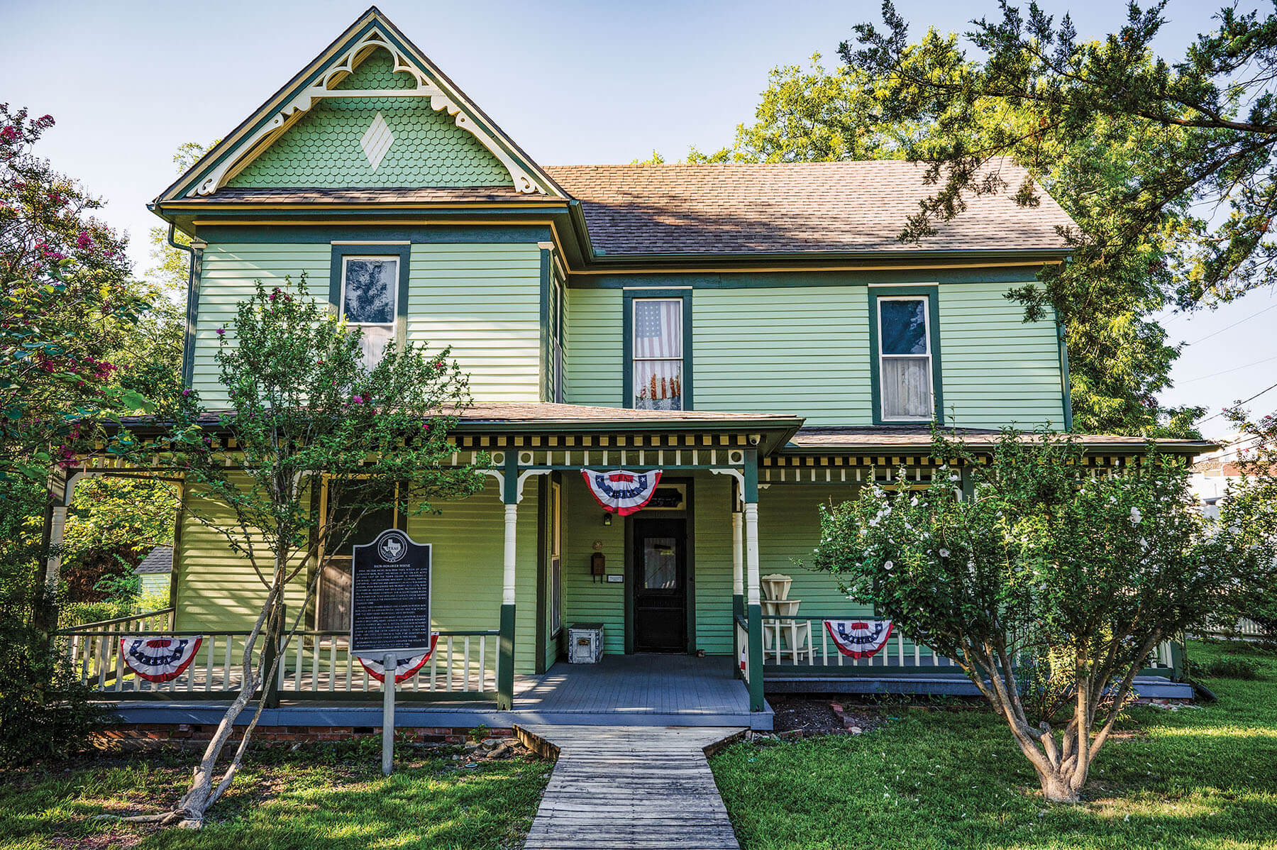 The exterior of a green historic home with tall green trees and lush green grass