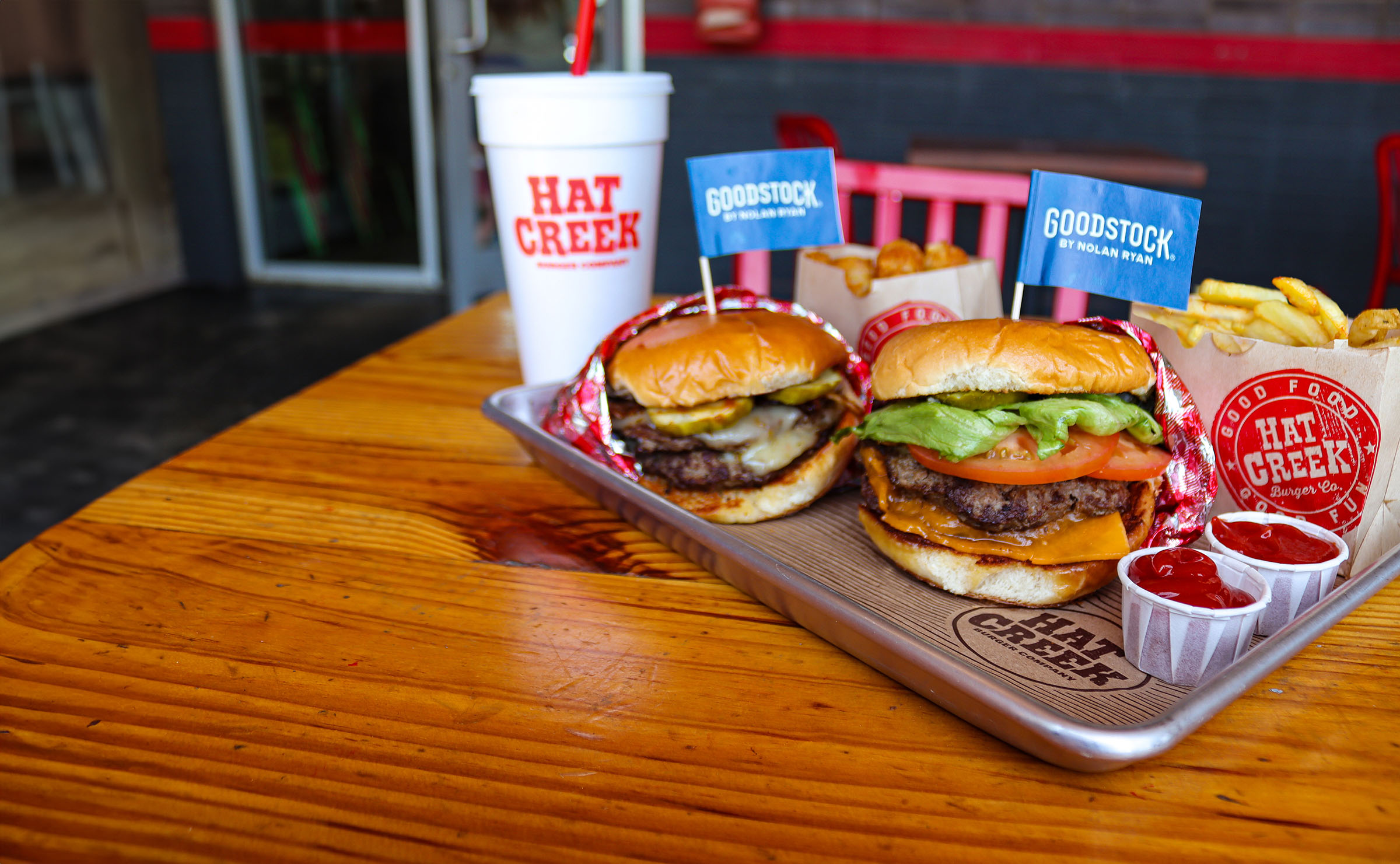 Two fully-loaded burgers with cheese and vegetables on a silver tray alongside small cups of ketchup, fries, and a drink in a Styrofoam cup