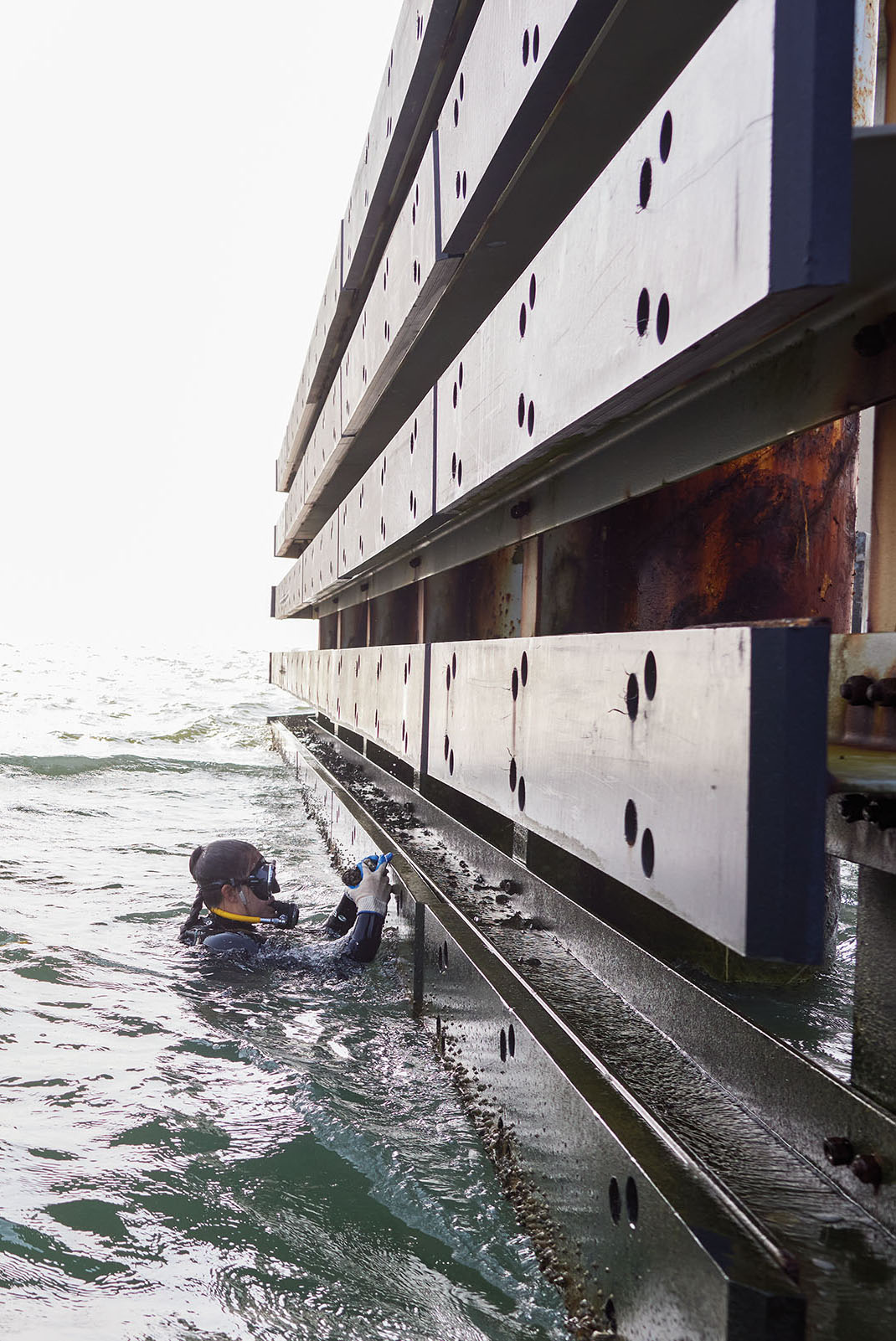 A person in SCUBA gear hangs off the side of a large structure in wavy water