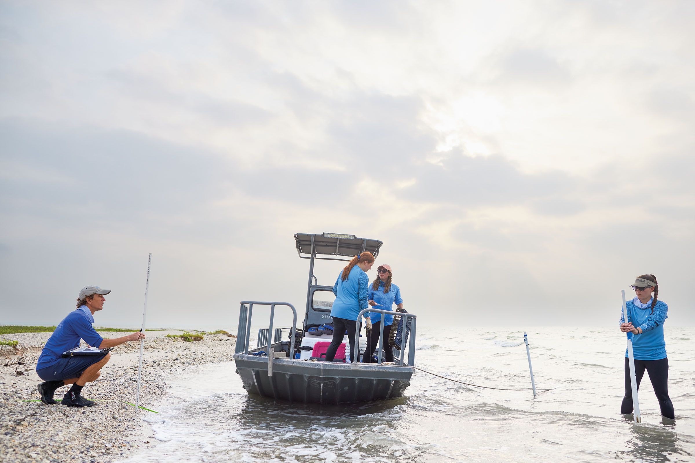 A group of people, some in a boat and some on shore, use scientific instruments in water