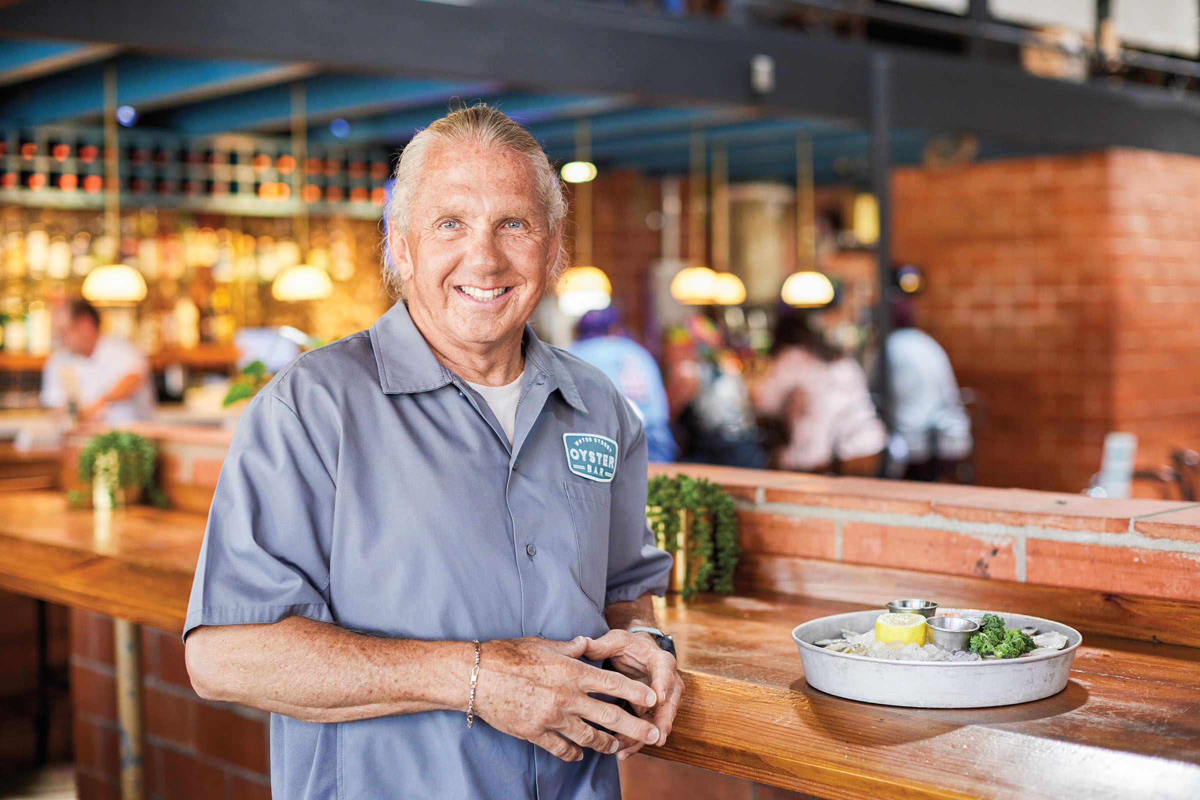 A man in a blue-grey shirt stands next to a wooden bar. A steel plate of oysters sits next to him