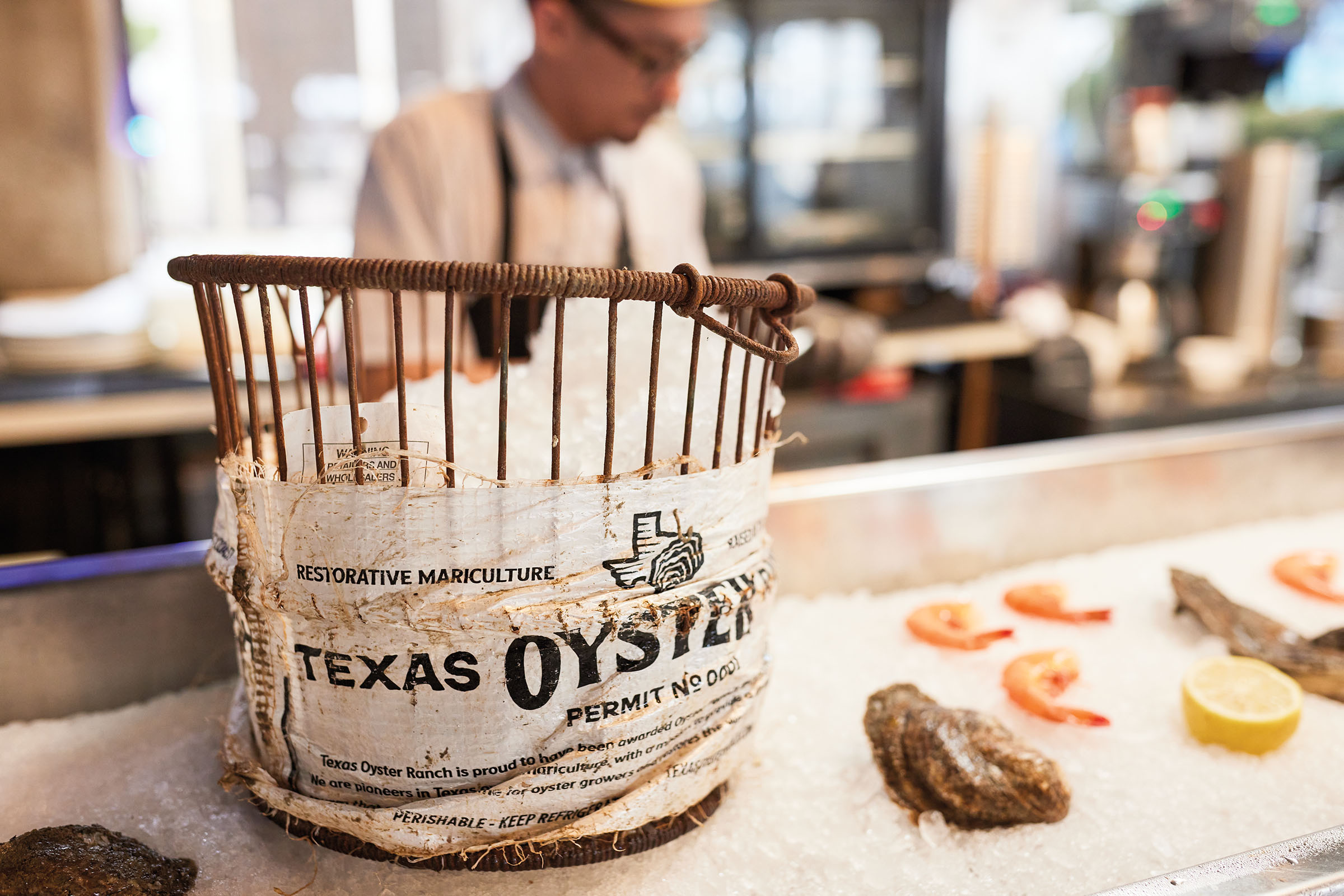 A small wooden basket with a wrapper reading "Texas Oysters" in the foreground, a man shucks oysters and works with other seafood in the background