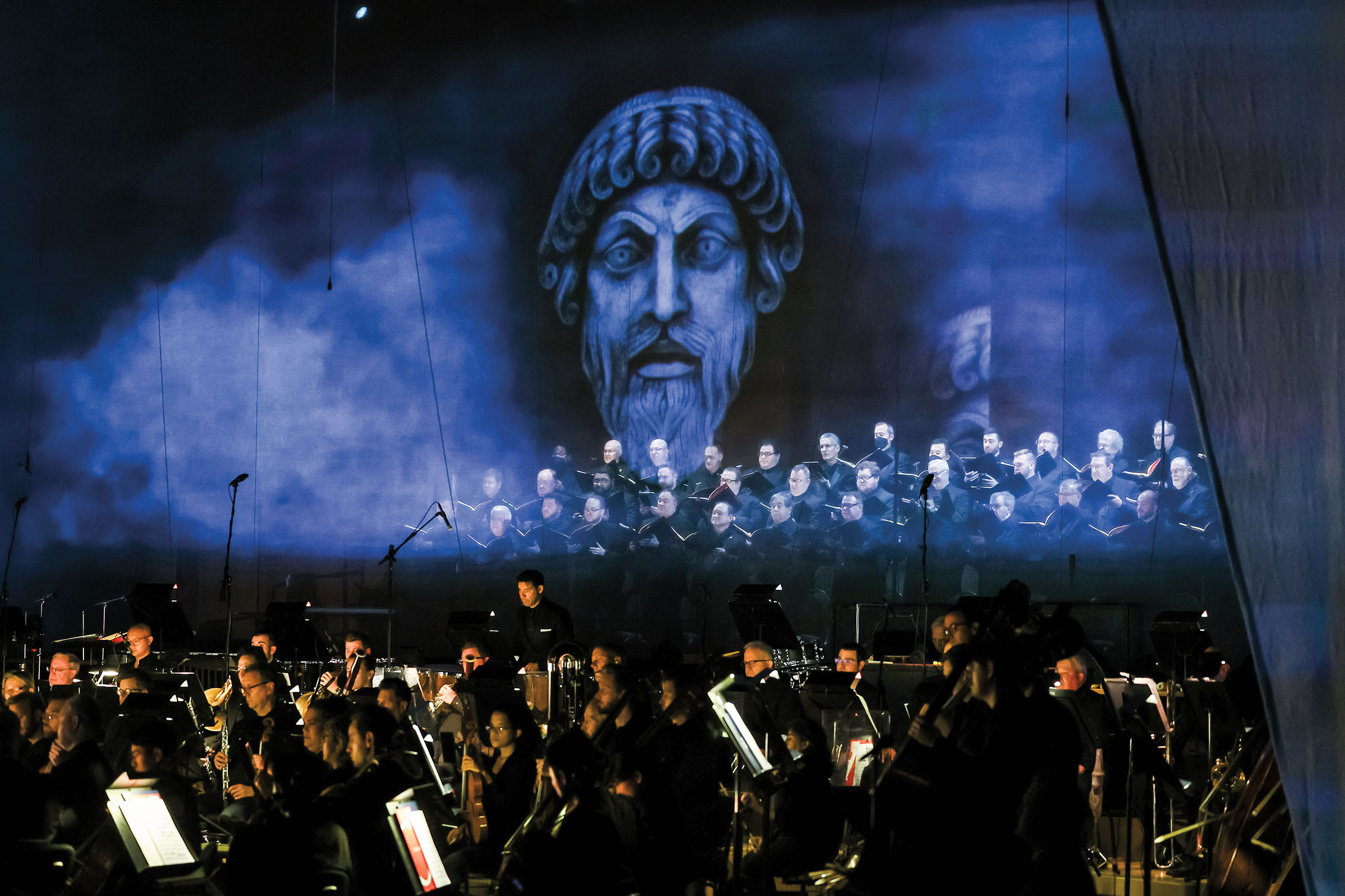 A large group of musicians play under a smoky blue projected image of a Roman sculpture